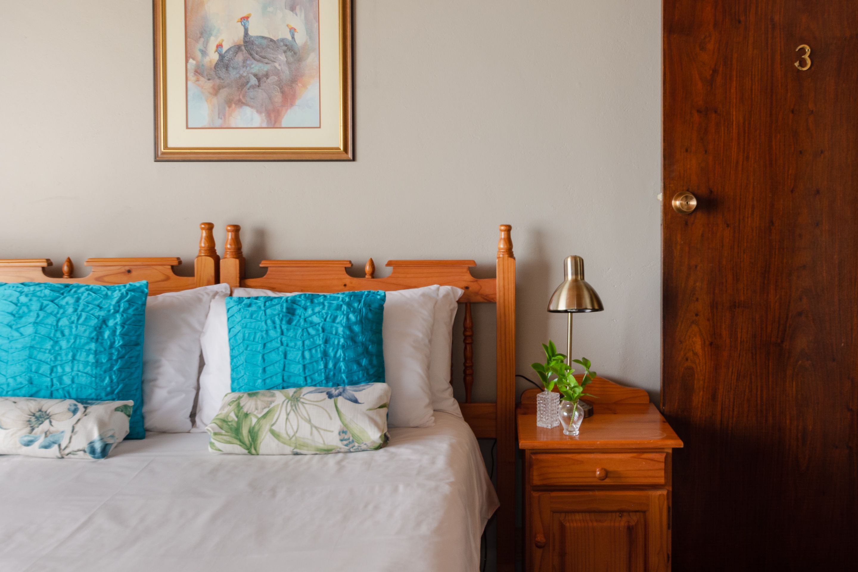 A neatly made bed with a white and blue floral pillow, a wooden headboard, and a bedside table with a lamp and a small plant.