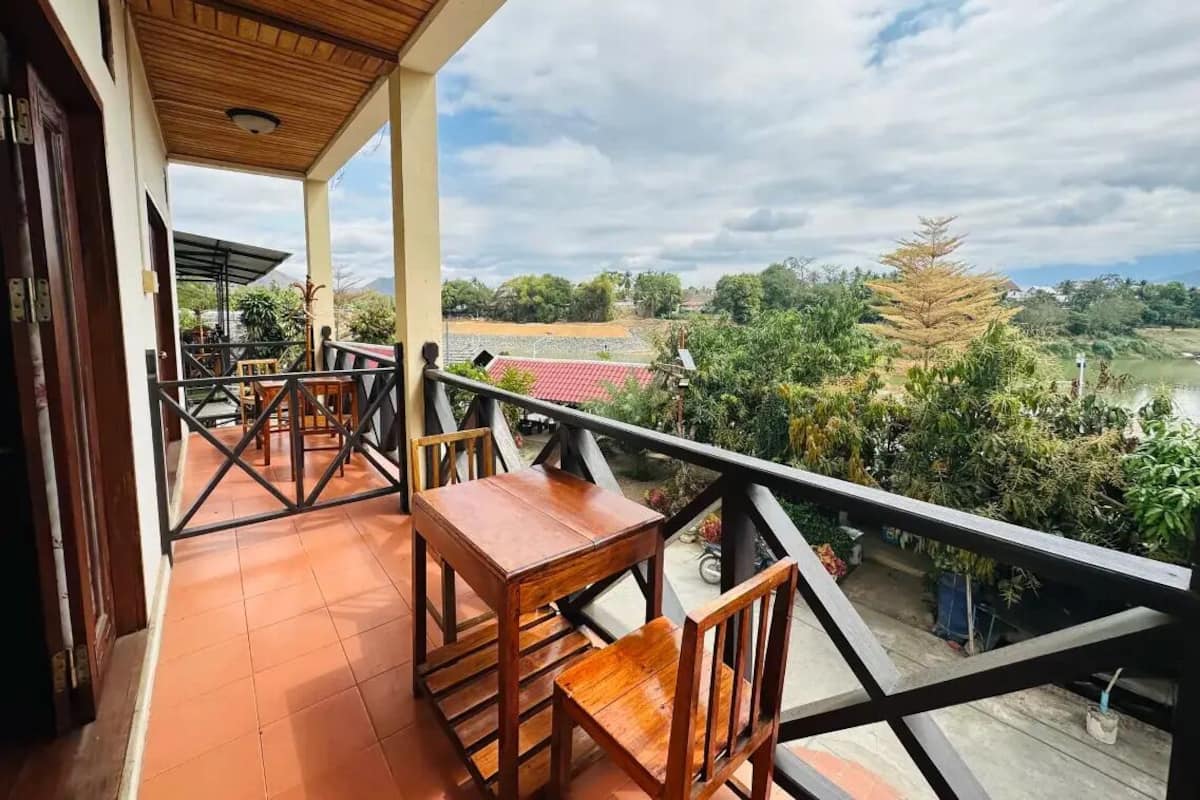 A balcony with a wooden table and chairs, a view of a rural landscape, and a red-roofed building in the distance.