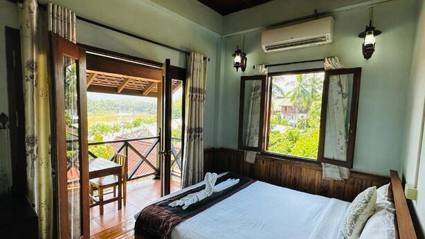 A bedroom with a bed, a window, an air conditioning unit, and a balcony with a table and chair. - Villa Kingkham Riverside (Luang Prabang)