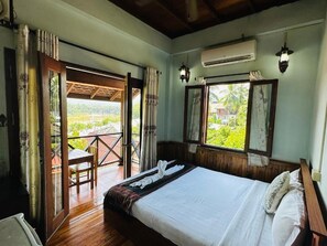 A bedroom with a bed, a window, an air conditioning unit, and a balcony with a table and chair. - Villa Kingkham Riverside (Luang Prabang)