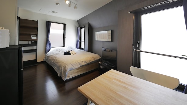 A modern bedroom with a bed, a desk, and a chair. There is a window with curtains, a flat-screen TV, and a wall-mounted shelf. - Apartment Sun Bright (Tokyo)