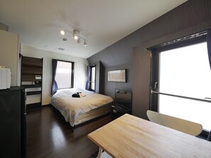 Un dormitorio moderno con cama, escritorio y silla. Hay una ventana con cortinas, un televisor de pantalla plana y un estante fijado a la pared.