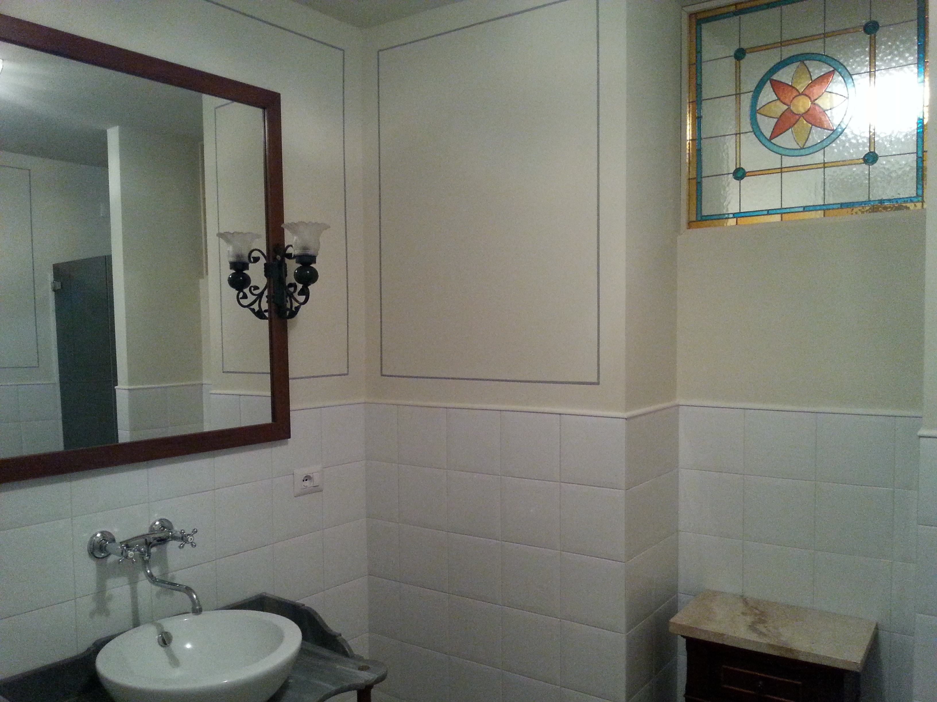 Un baño con paredes de azulejos blancos, un espejo grande, un lavabo y un ventanal con vidrieras de colores.