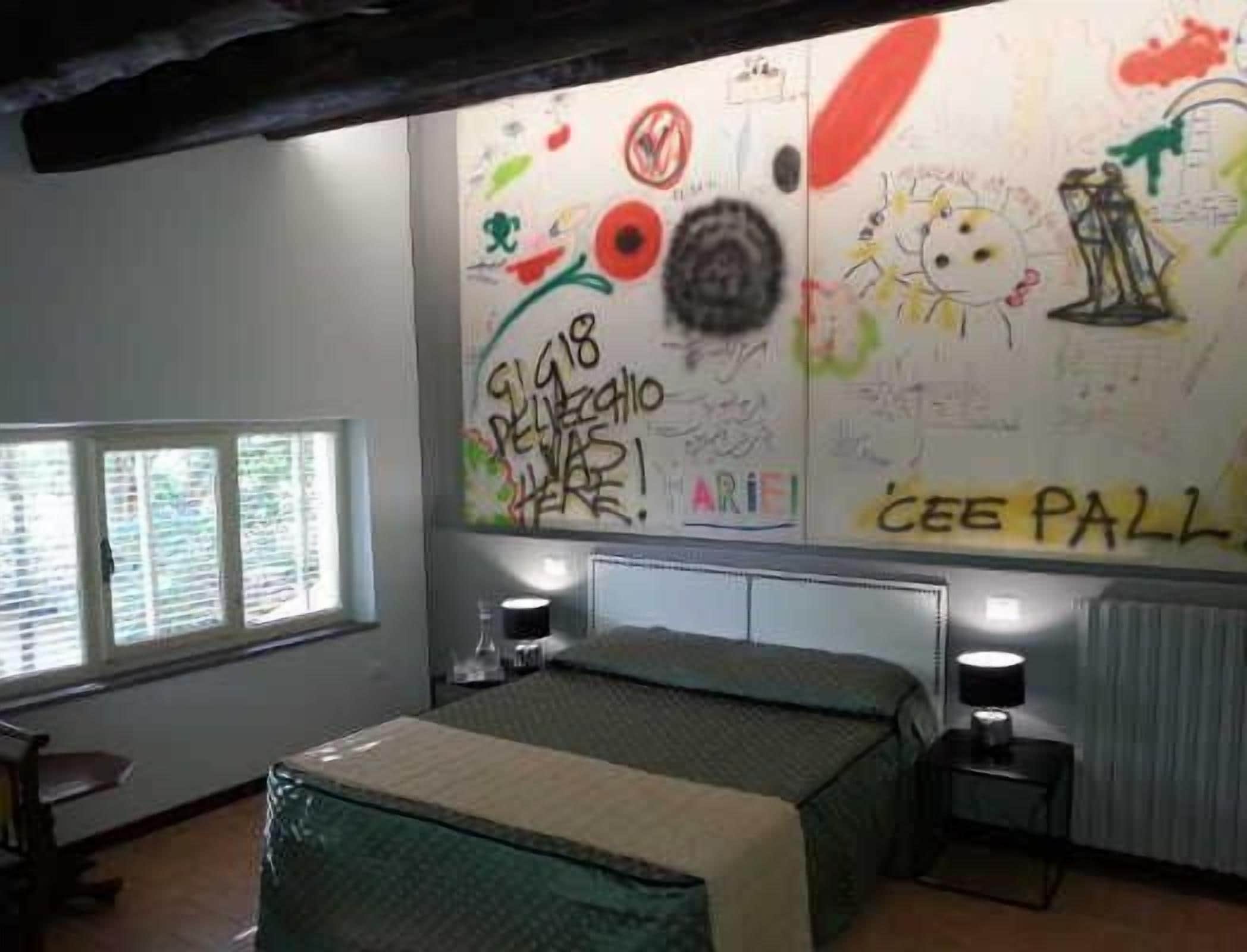 Una habitación con una cama, mesitas de noche, un ventanal con persianas y una pared con grafitis y dibujos.