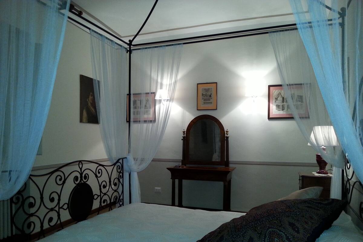 Un dormitorio con una cama con dosel, una mesita de noche con lámpara y un espejo en la pared.