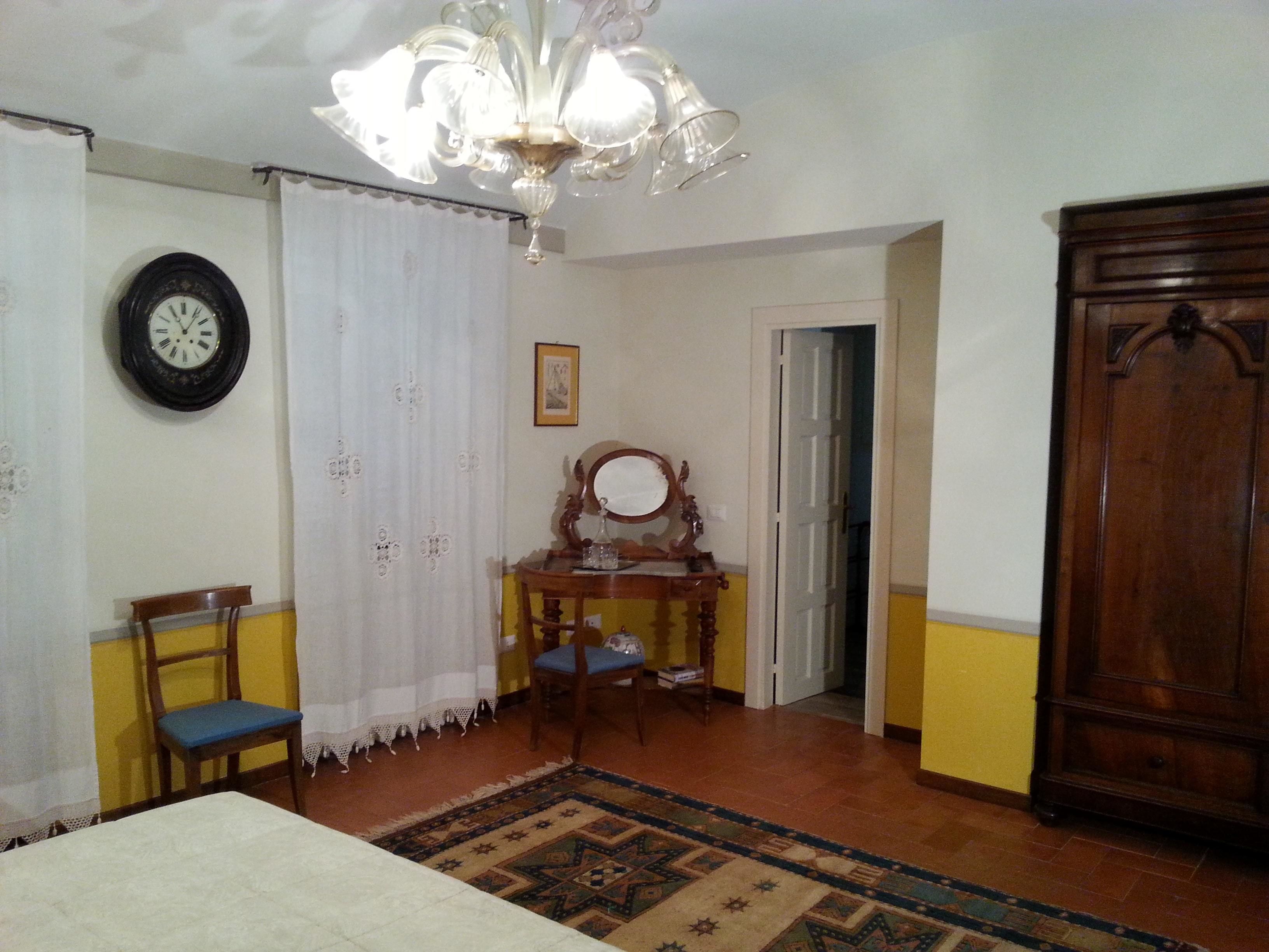 A room with a bed, a wooden chair, a table with a mirror, a clock on the wall, and a door leading to another room.