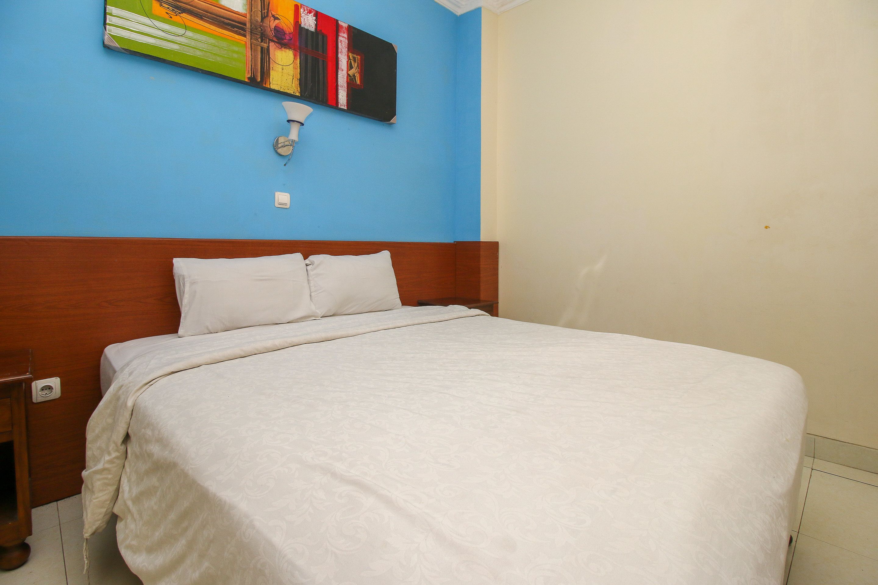 A bedroom with a bed, a nightstand, a wall-mounted light, and a colorful abstract painting.