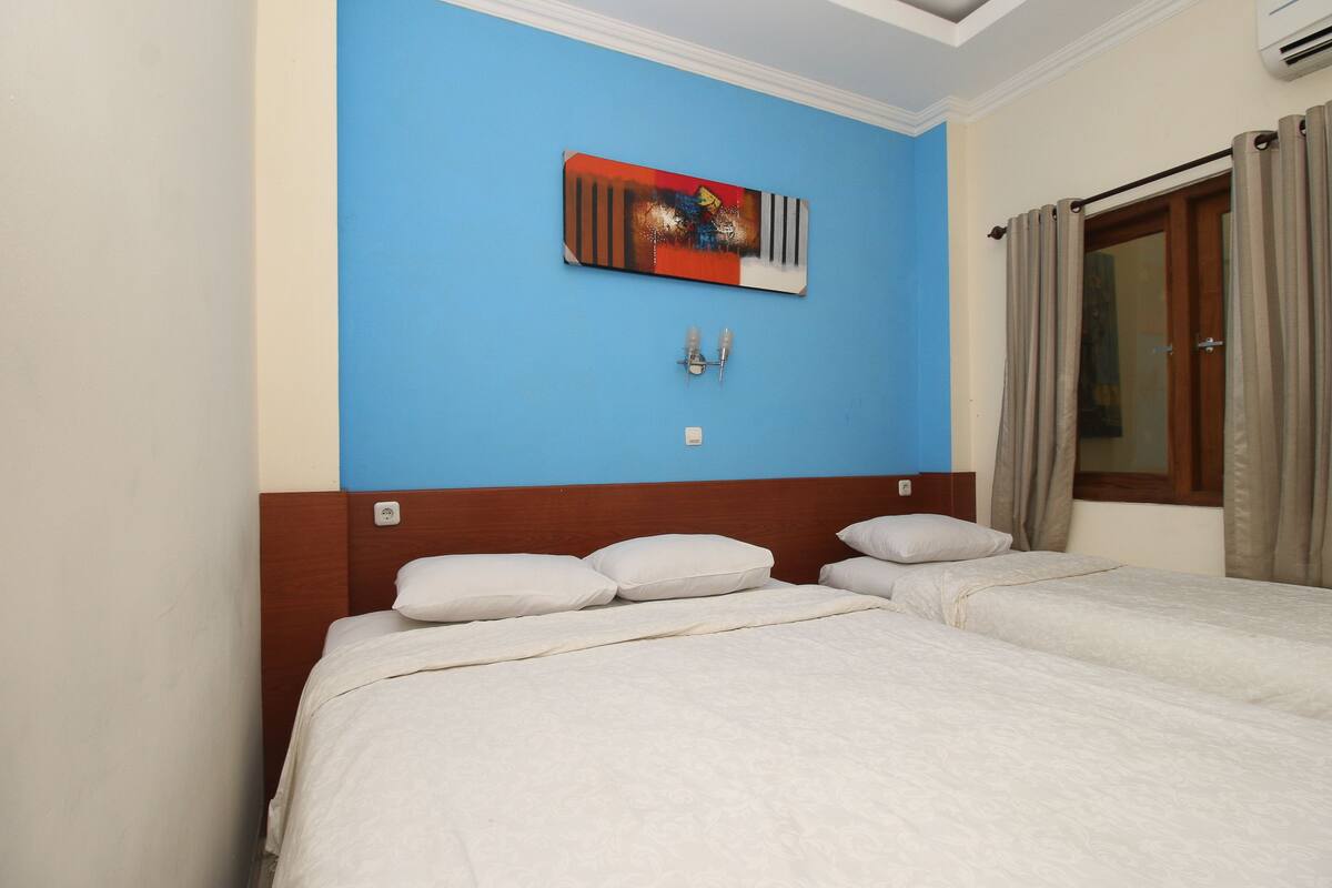 A hotel room with two beds, a blue accent wall, a decorative piece above the beds, and a window with curtains.