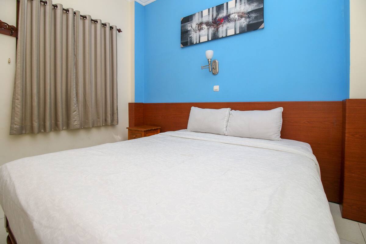 A bedroom with a large bed, a nightstand, a wall-mounted light, and a window with curtains.