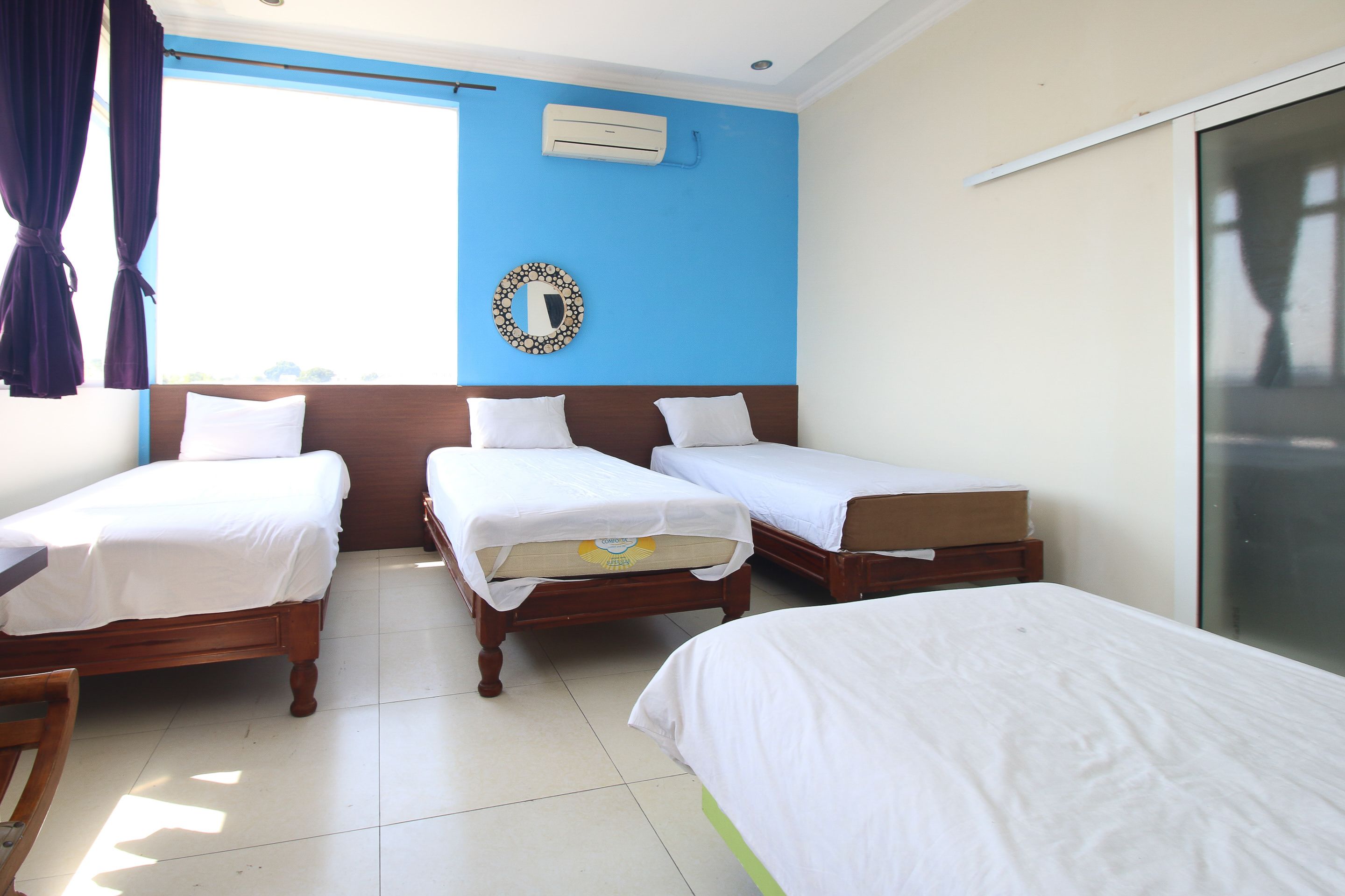 A hotel room with three single beds, a large window, an air conditioning unit, and a mirror on the wall.