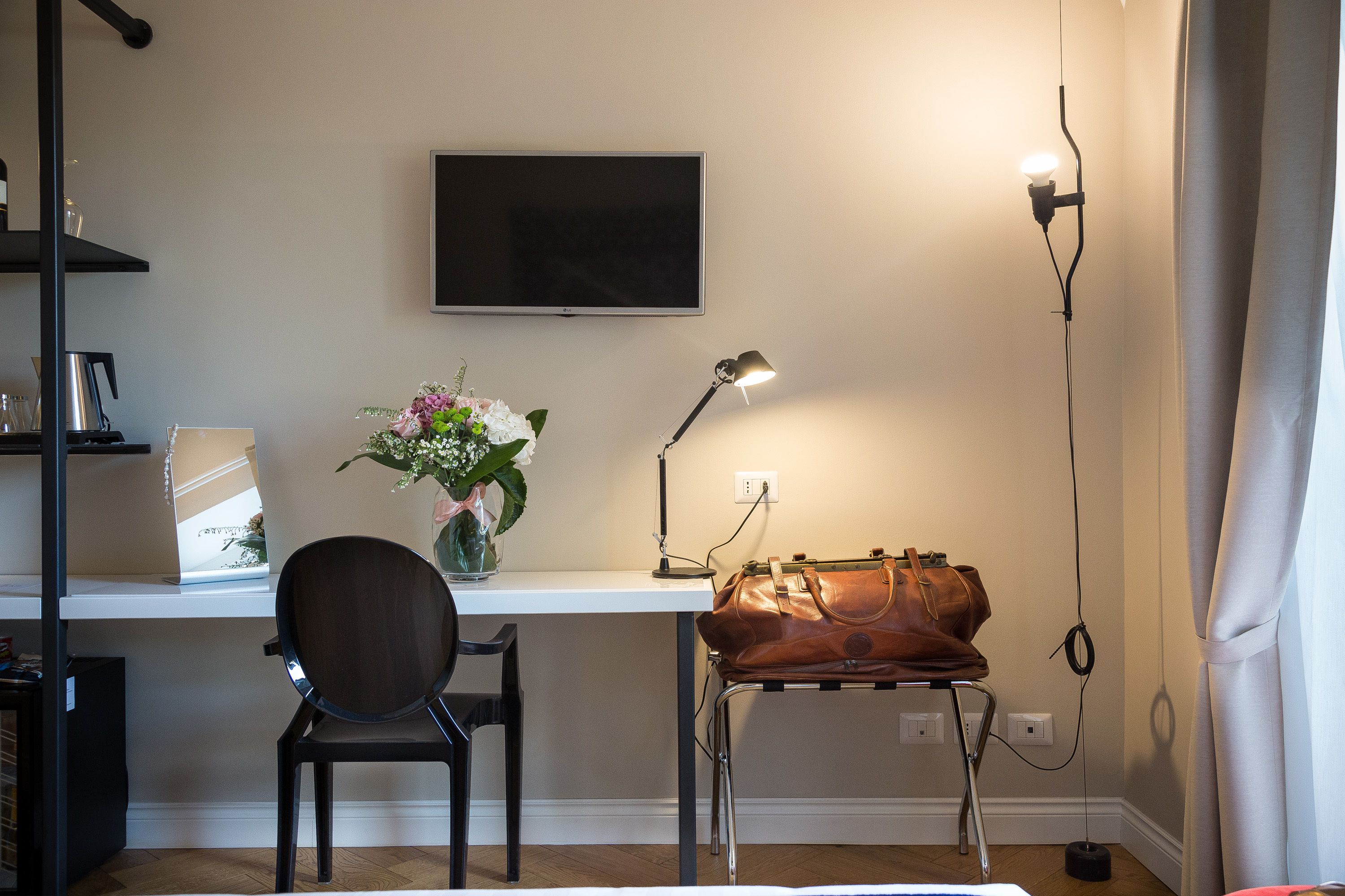 Ett rum med ett skrivbord, en stol, en tv som är monterad på väggen, en lampa, en vas med blommor och en väska på skrivbordet.