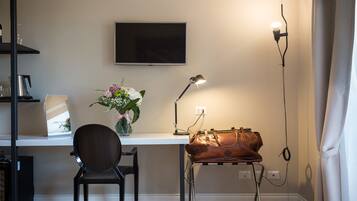 Ett rum med ett skrivbord, en stol, en tv som är monterad på väggen, en lampa, en vas med blommor och en väska på skrivbordet.