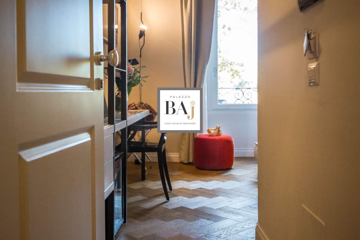 A hallway with a wooden herringbone floor, a red ottoman, a black chair, and a table with a vase of flowers.