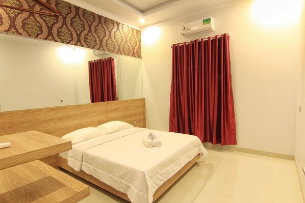 Une chambre d’hôtel avec une tête de lit en bois, un lit avec une literie blanche, une table de chevet en bois, un miroir, des rideaux rouges et un appareil de climatisation.