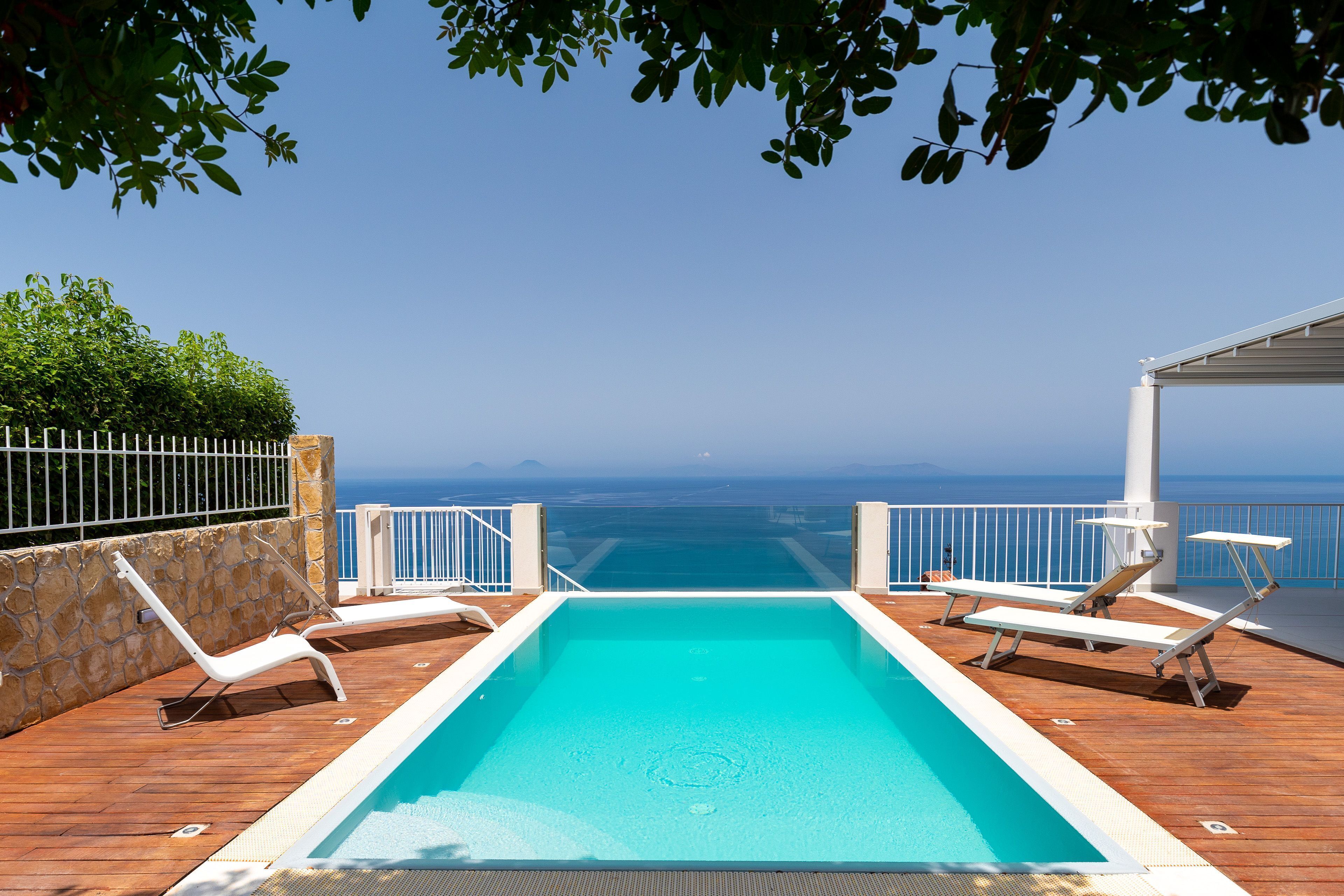 A pool area with a view of the ocean, surrounded by a wooden deck and white railings.