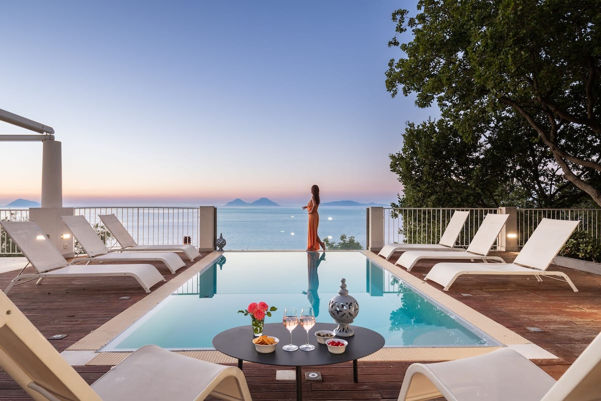 A poolside area with lounge chairs and a table set for two, overlooking a body of water and distant mountains.