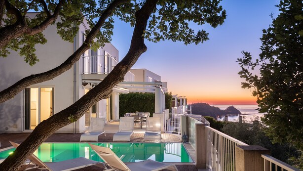 A modern outdoor pool area with lounge chairs, a dining table, and a view of the ocean at sunset.