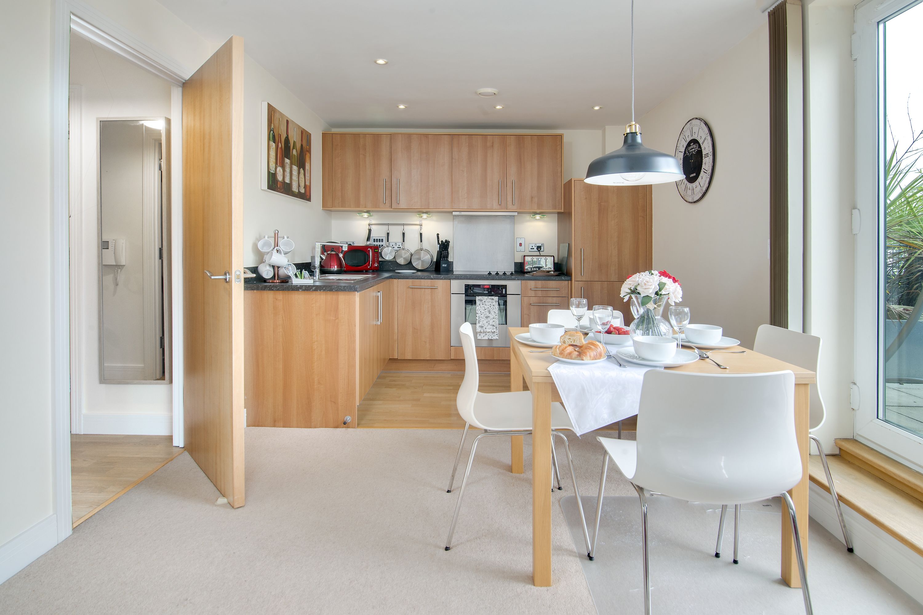 Une cuisine moderne avec des meubles en bois, une table à manger entourée de chaises blanches et une grande fenêtre qui laisse entrer la lumière naturelle.