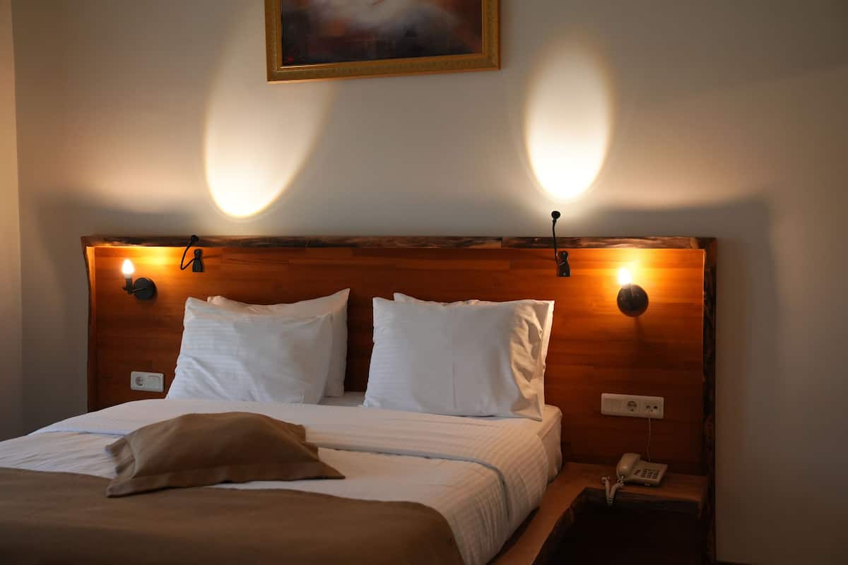 A neatly made bed with white linens and a brown throw pillow, a wooden headboard with wall-mounted reading lights, and a bedside telephone.