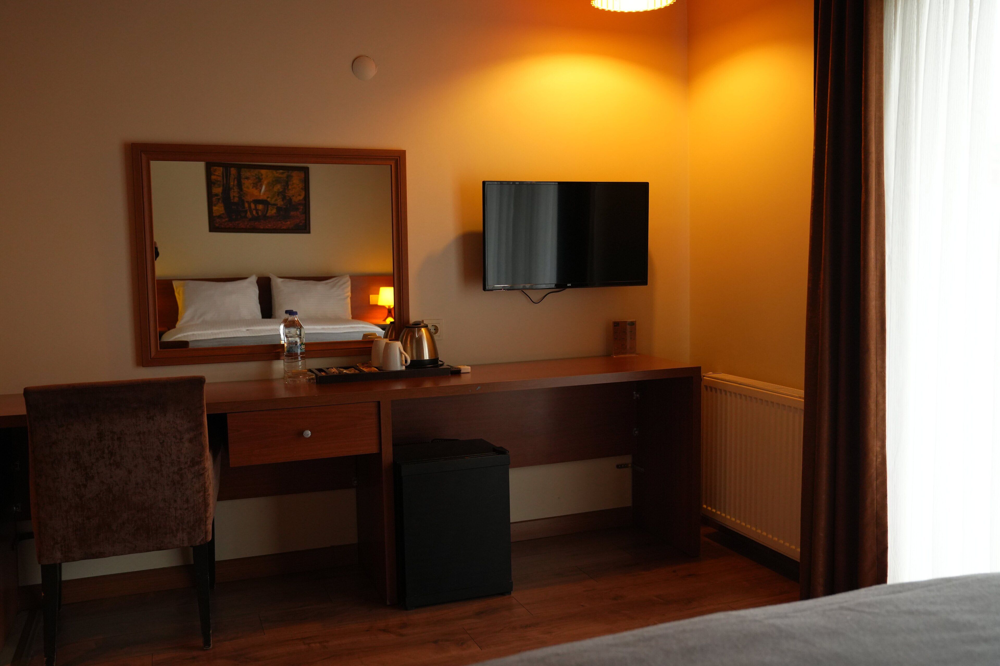Ein Hotelzimmer mit einem Holztisch, einem Stuhl, einem Bett, einem an der Wand montierten Fernseher und einem Fenster mit Vorhängen.