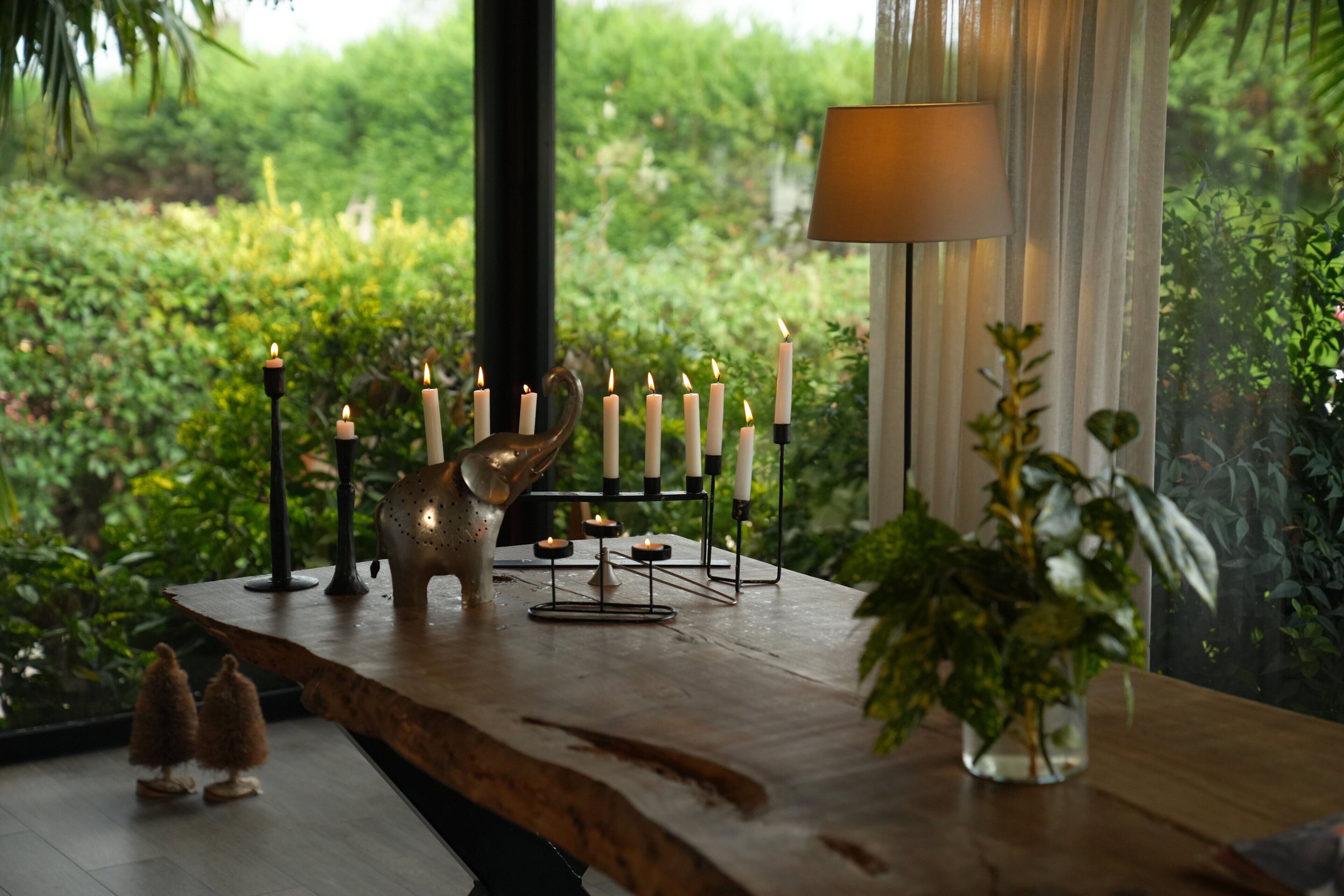 A wooden table with candles and a lamp, surrounded by greenery.