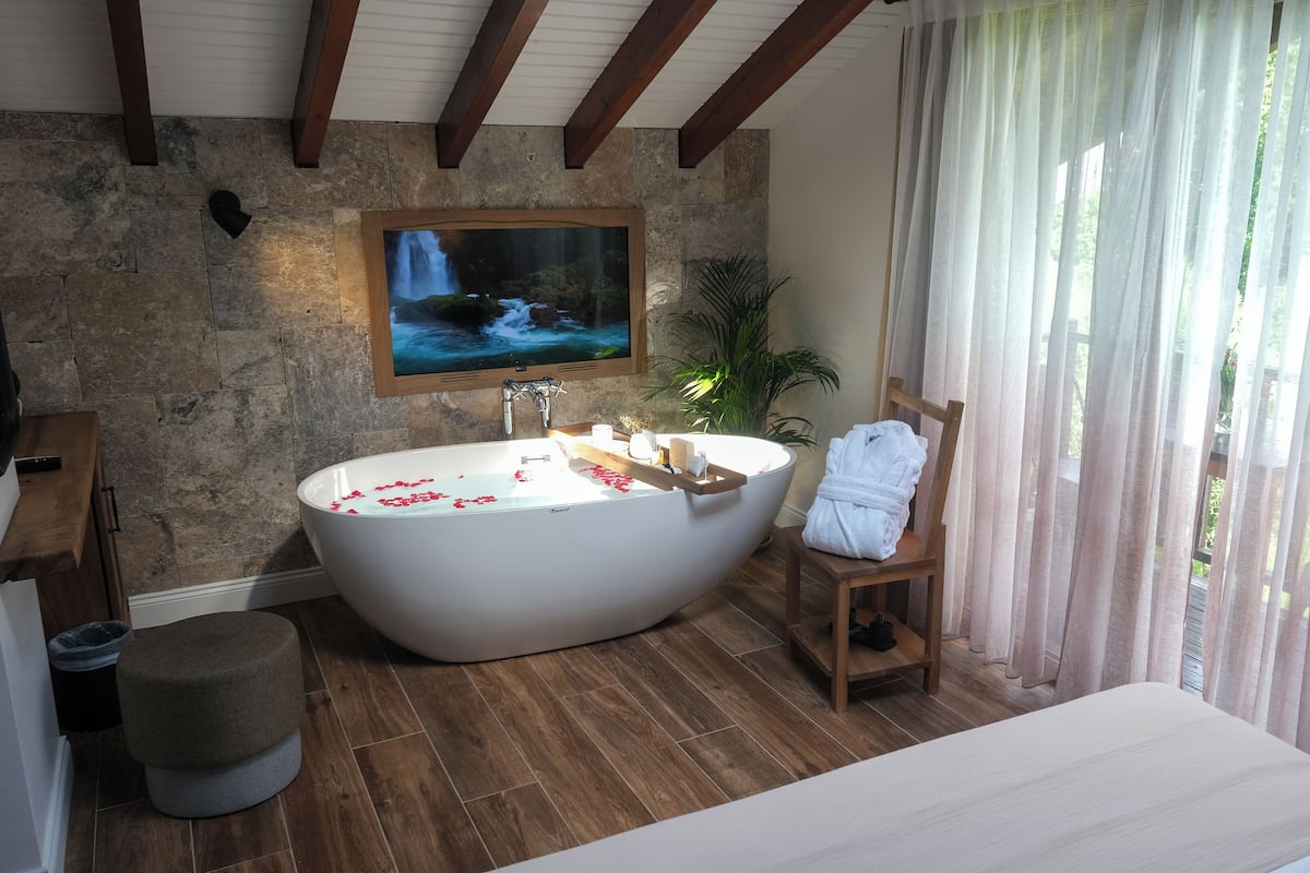 A modern bathroom with a freestanding bathtub, a wooden bench, a large mirror, and a stone wall.