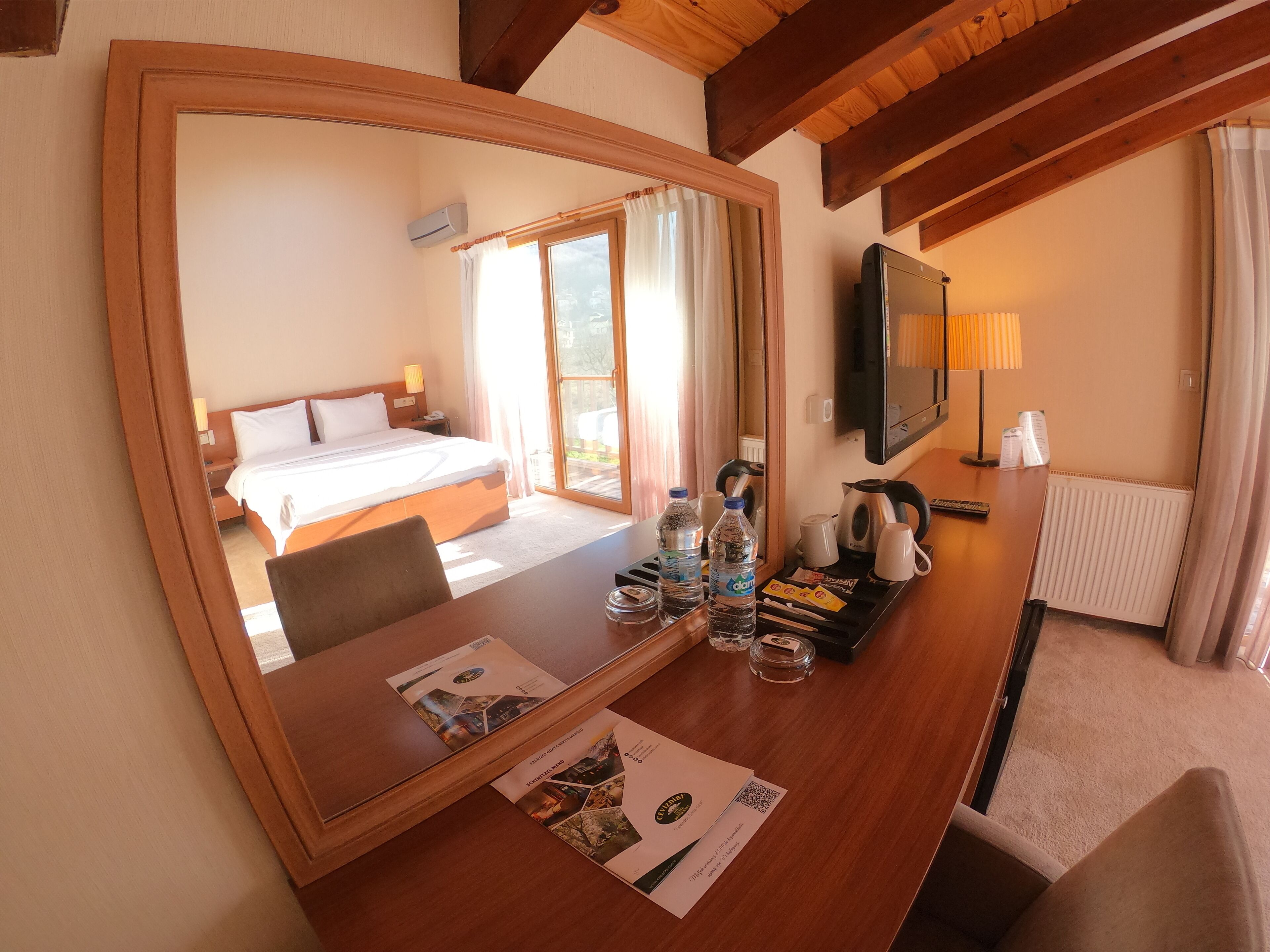A hotel room with a large mirror, a bed, a desk with a laptop, a TV, and a window with curtains.