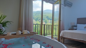 A bedroom with a bed, a bedside table with a lamp, a window with curtains, a view of greenery and houses, and a bathtub with rose petals. - Cevizdibi Otel (Kartepe)
