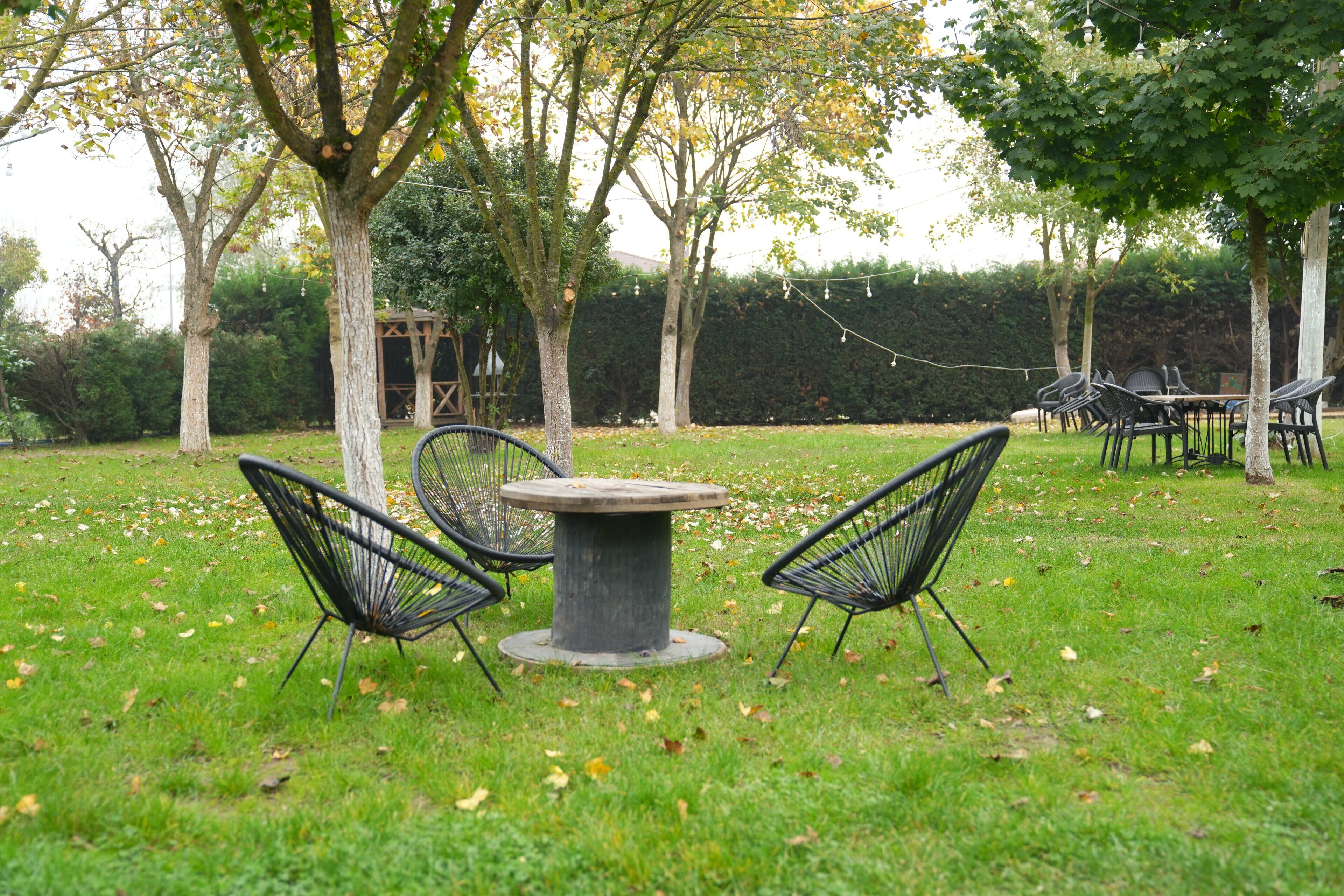 An outdoor seating area with a round table and four chairs, surrounded by trees and a hedge in the background.