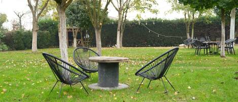 An outdoor seating area with a round table and four chairs, surrounded by trees and a hedge in the background.