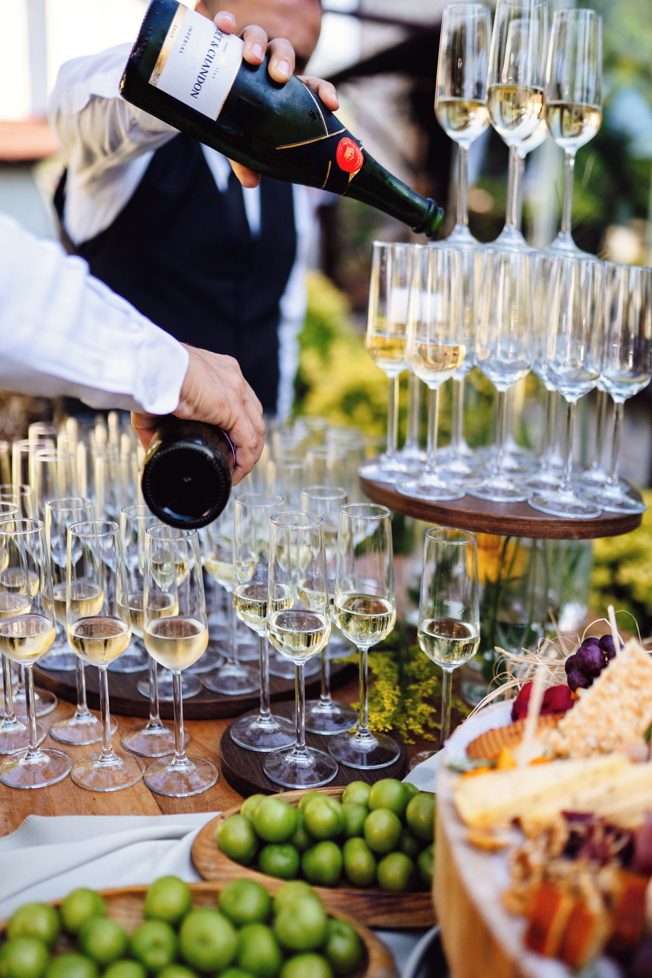 A person pouring wine into glasses, a cheese board with various cheeses, and a wooden table with wine glasses and a bowl of green grapes.