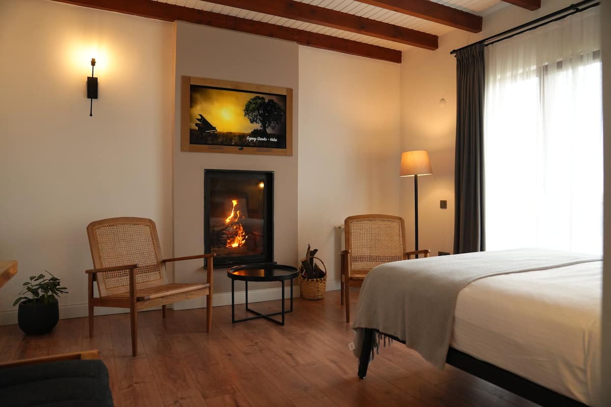 A hotel room with a fireplace, two wicker chairs, a bed, a nightstand, and a lamp.