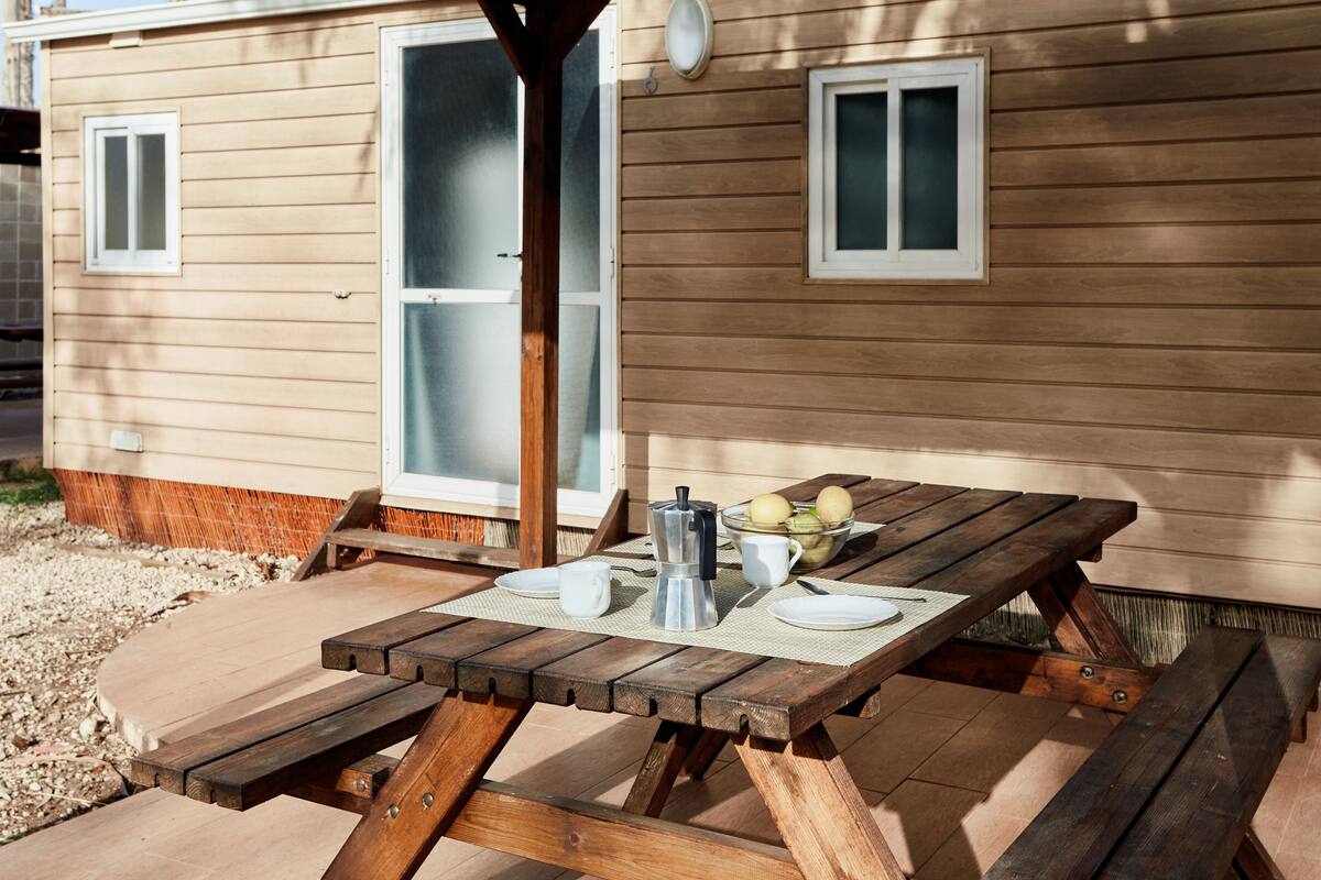 Ein Holztisch für ein Picknick mit einer Metall-Kaffeekanne, weißen Tassen und einer Schüssel mit Obst.