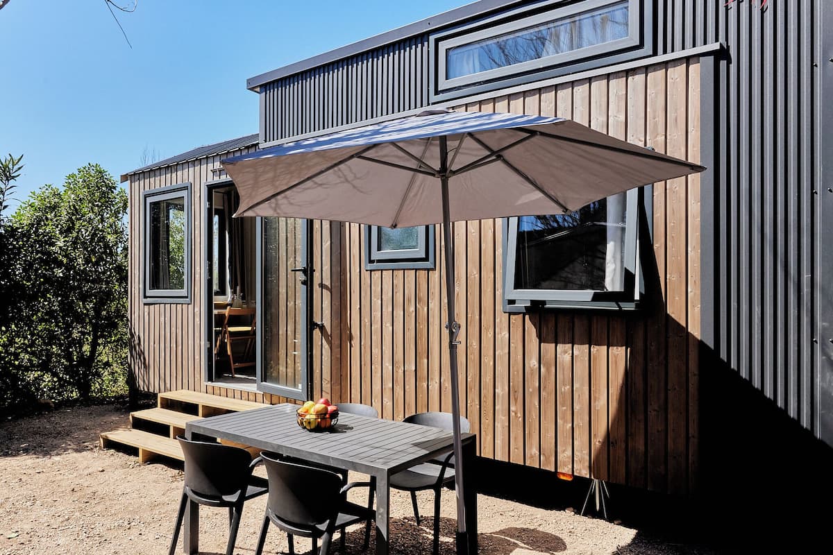 Eine Holzhütte mit Essbereich, inklusive Tisch, Stühlen und einem großen Sonnenschirm sowie einem kleinen Fenster.