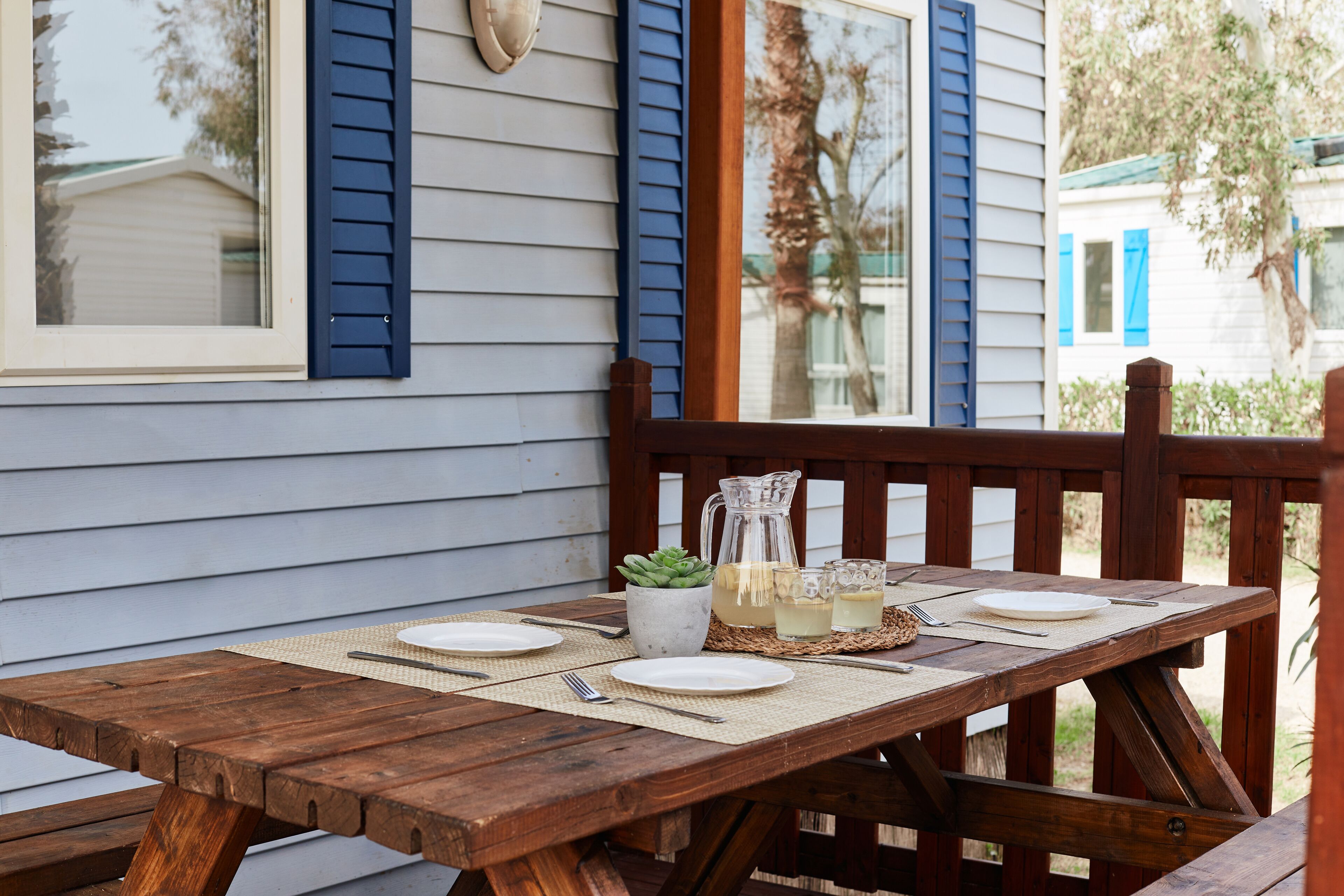 Un tavolo da pranzo in legno all'aperto apparecchiato con piatti, posate e una brocca di limonata, posizionato su un portico con vista su una casa con persiane blu.