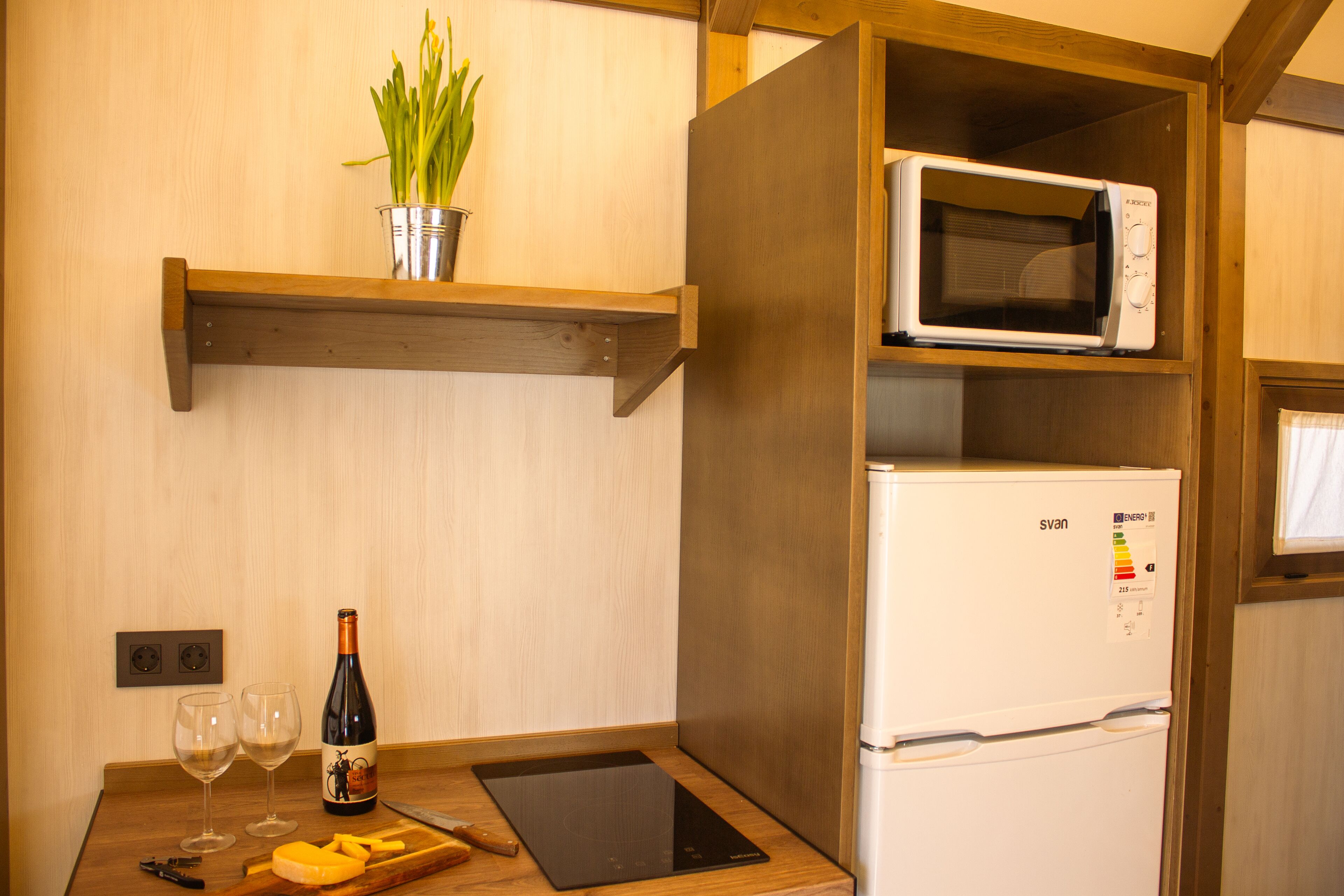 Una cocina con microondas, refrigerador, estante de madera con una planta, botella de vino, vasos y tabla de cortar.