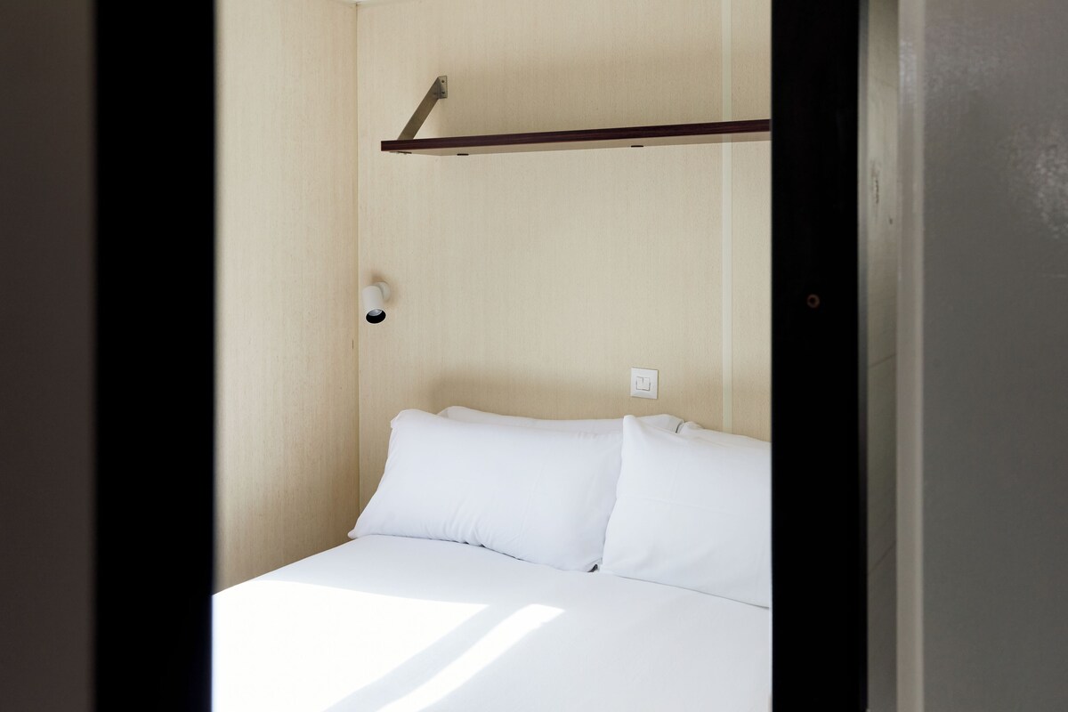 Una habitación pequeña con una cama individual, cabecero de madera, mesita de noche y un interruptor de luz en la pared.