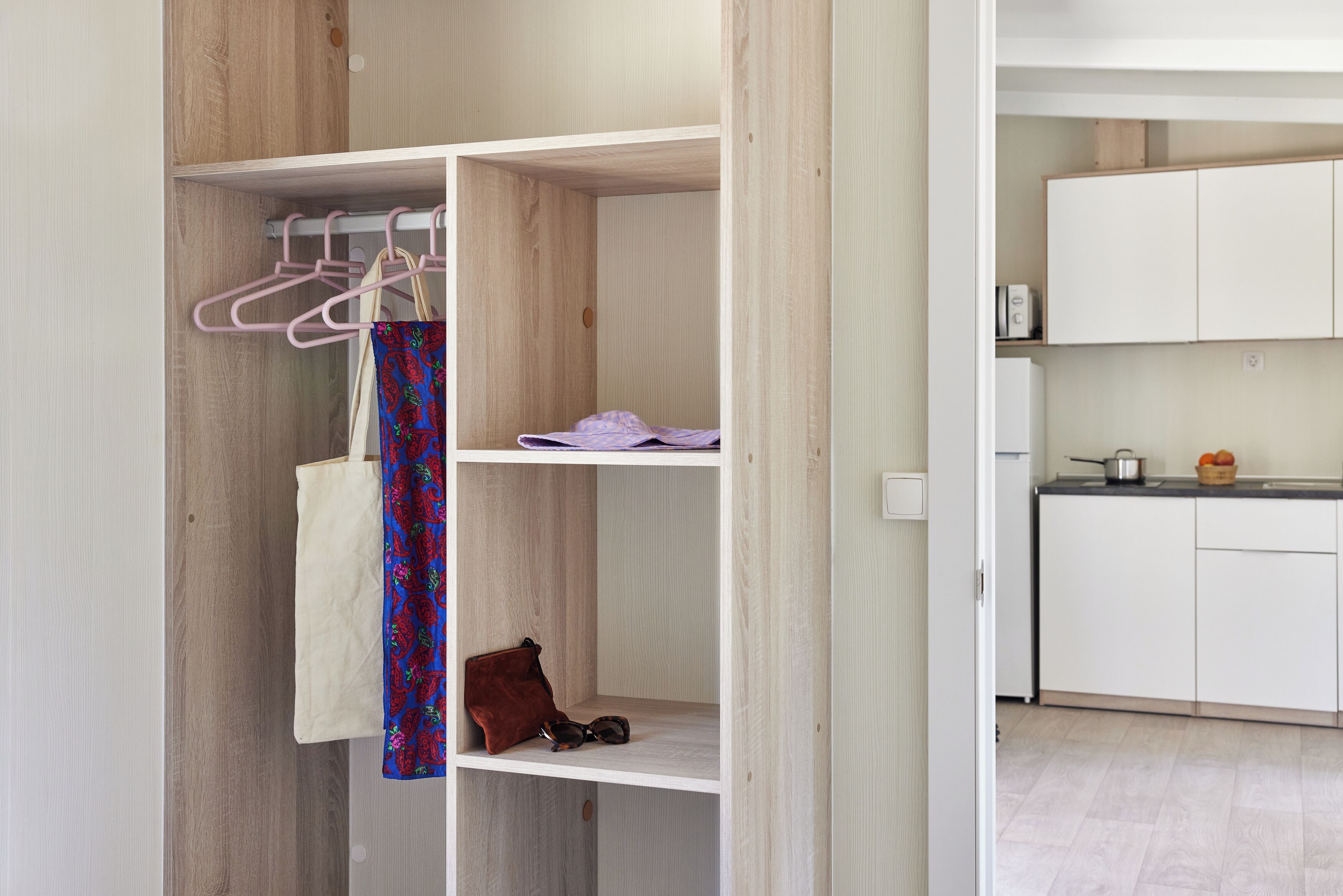 A modern, open-plan living space with a wooden wardrobe, a kitchen area, and a hanging area with clothes and a handbag.