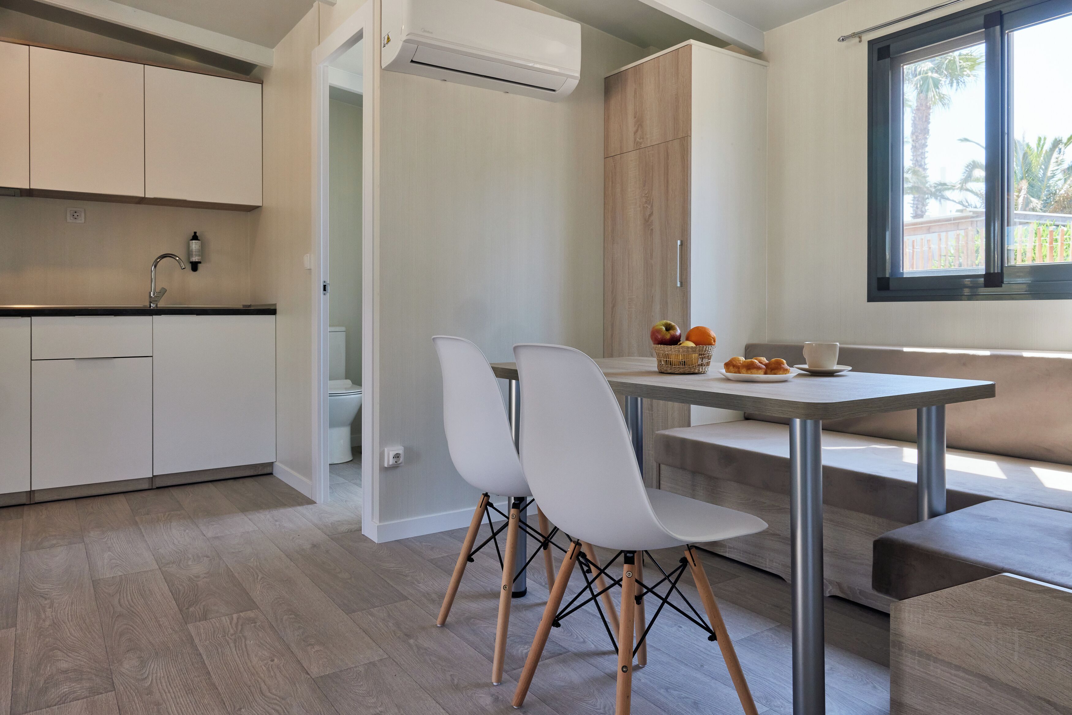 Een moderne keuken met witte kasten, een eethoek met een tafel en stoelen, en een raam met uitzicht op palmbomen.