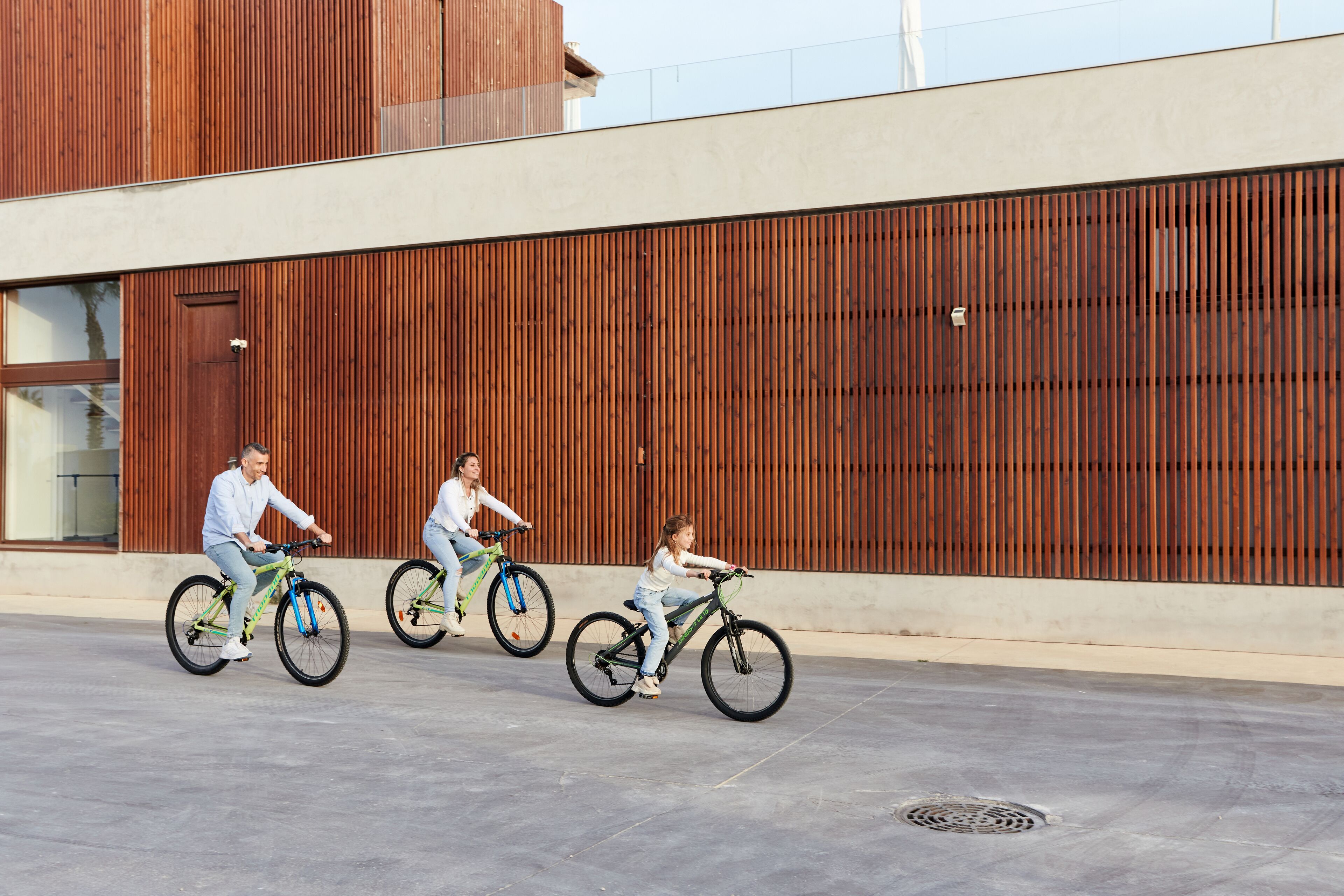 Três pessoas andando de bicicleta em uma superfície plana, com um edifício moderno ao fundo, caracterizado por ripas verticais de madeira.