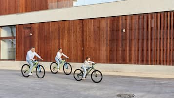 Três pessoas andando de bicicleta em uma superfície plana, com um edifício moderno ao fundo, caracterizado por ripas verticais de madeira.