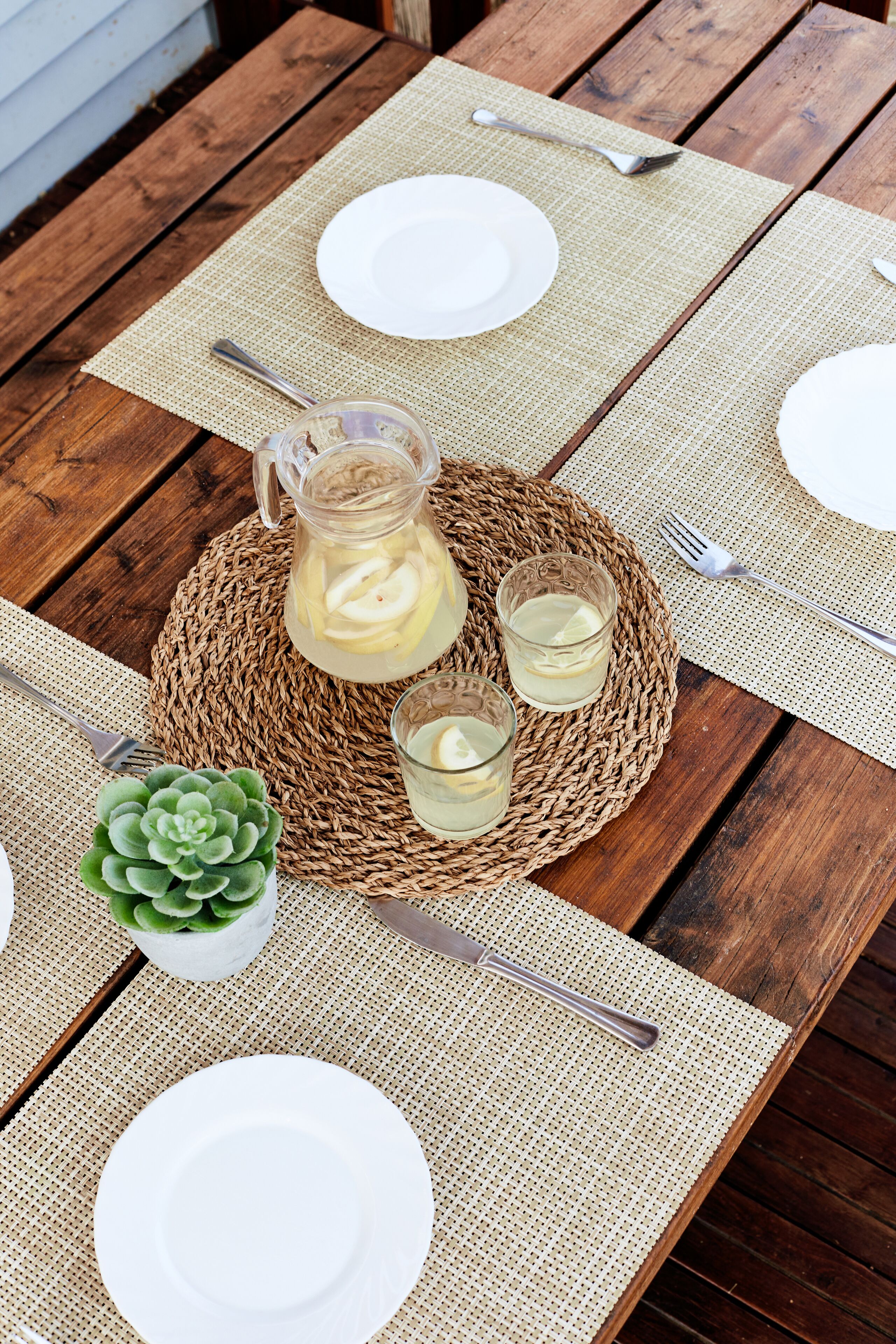 Uma mesa de madeira posta com pratos brancos, uma jarra de limonada e dois copos de limonada sobre um jogo americano de palha.