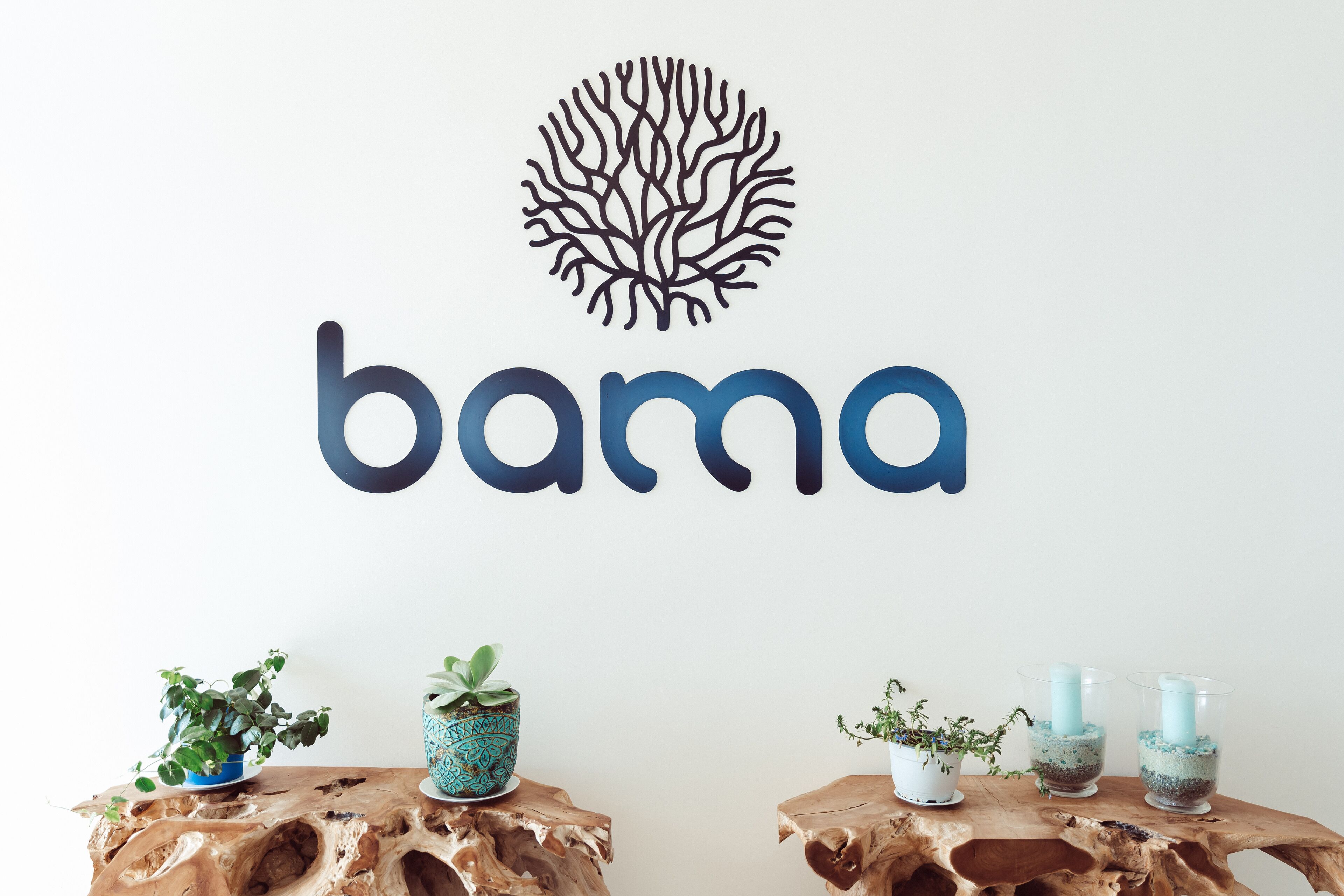 Eine Wand mit einem Logo und dem Schriftzug "bama" in großen Lettern. Zwei Holztische mit Topfpflanzen und einer blauen Vase sind zu sehen.