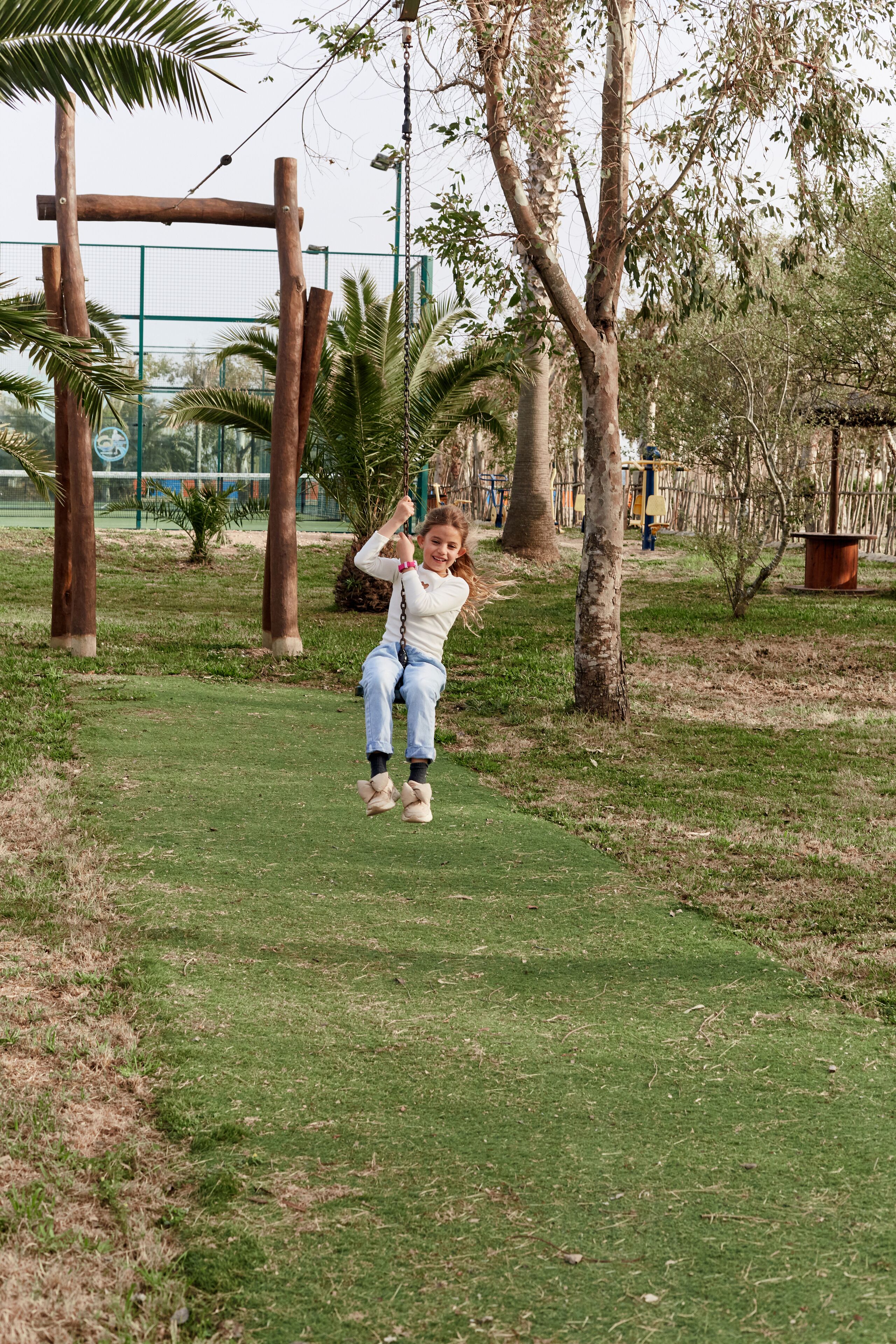 A child on a swing in a park with grass, trees, and a fence in the background.