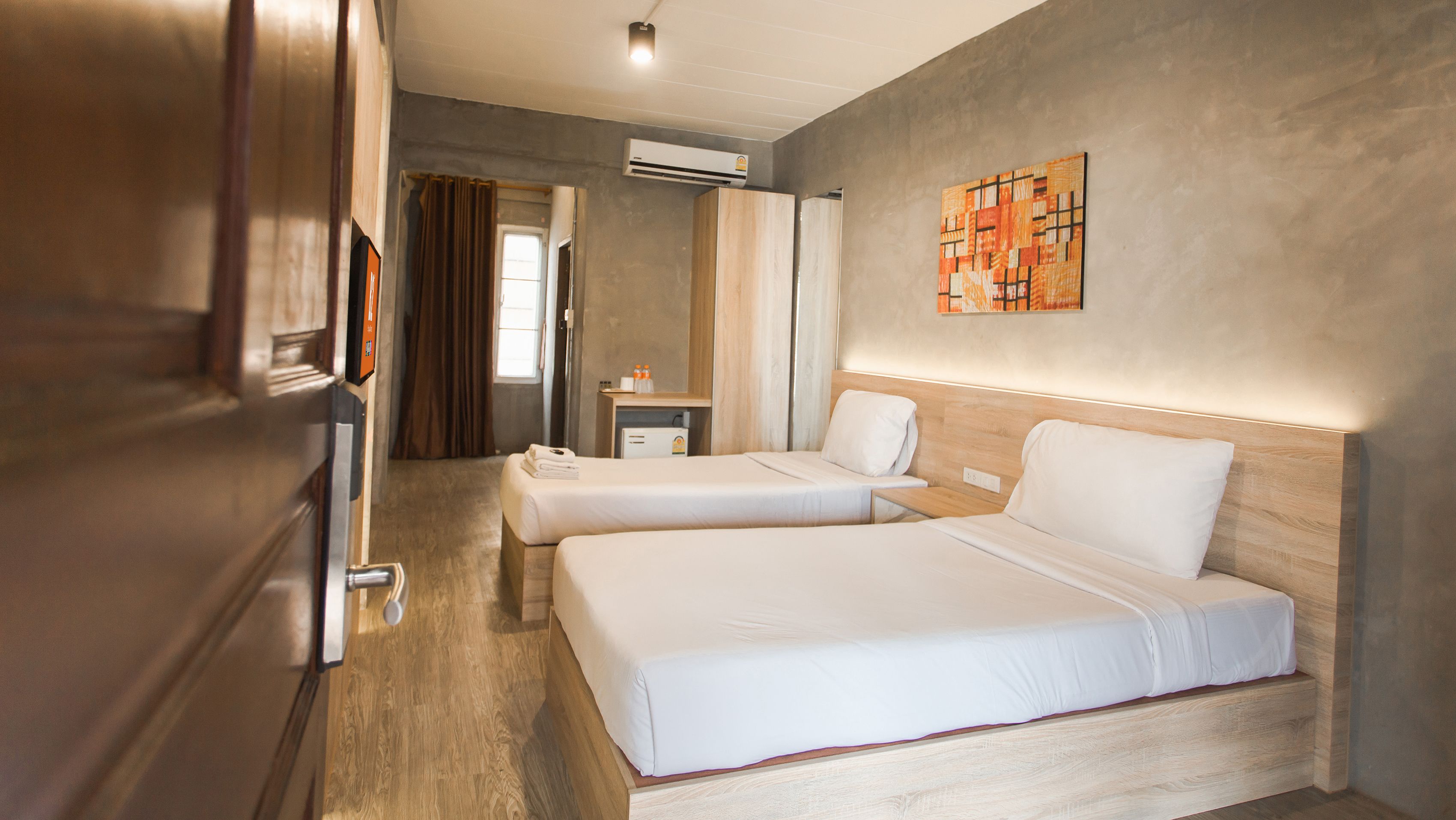 Habitación de hotel con dos camas, suelo de madera, unidad de aire acondicionado, mesita de noche pequeña y una obra de arte colgada en la pared.