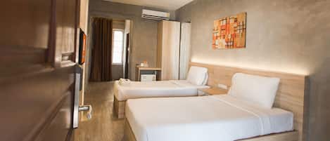 A hotel room with two beds, a wooden floor, an air conditioning unit, a small bedside table, and a wall-mounted artwork.