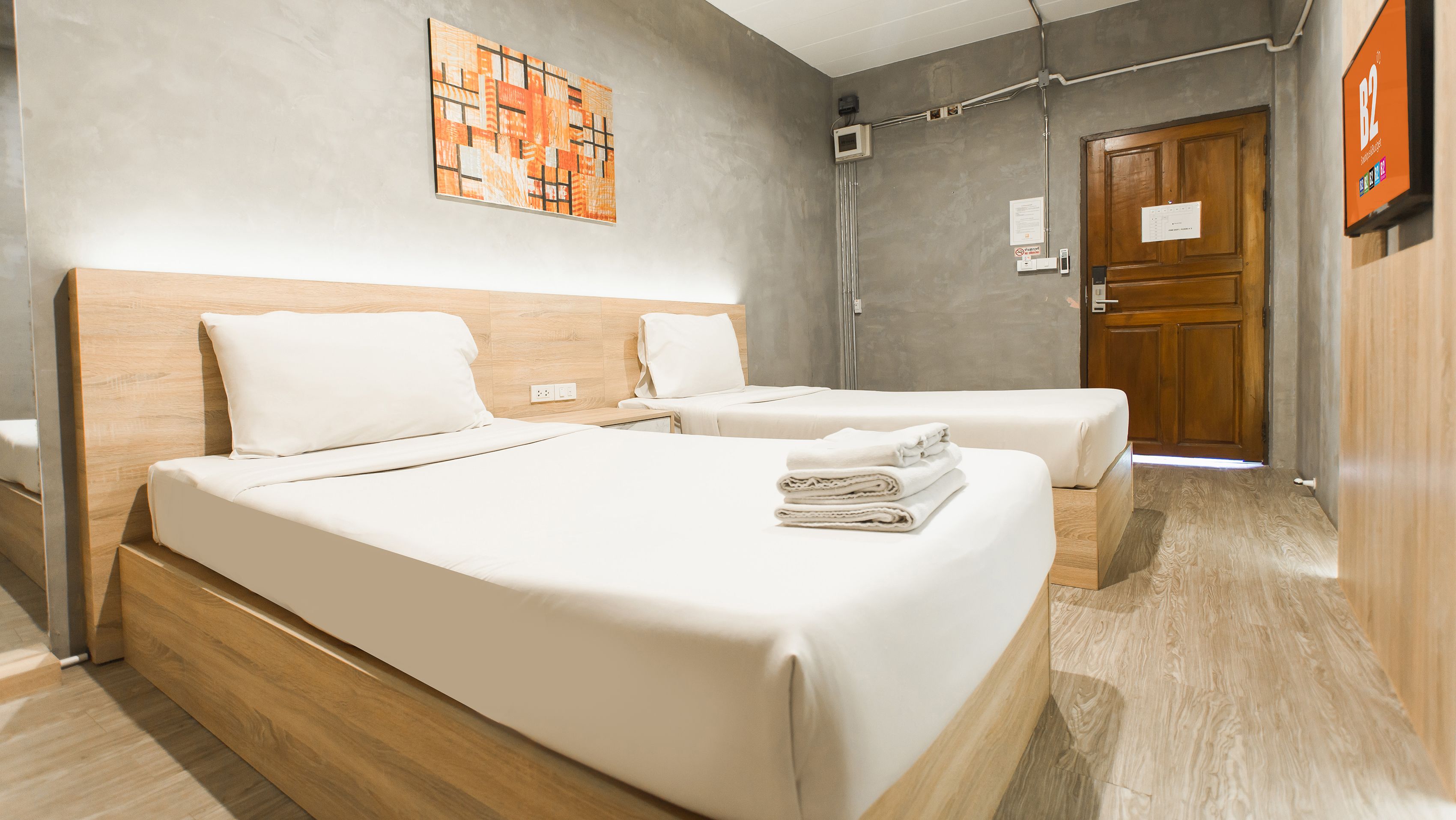 A hotel room with two beds, a wooden headboard, a wall art piece, a door, and a small table.