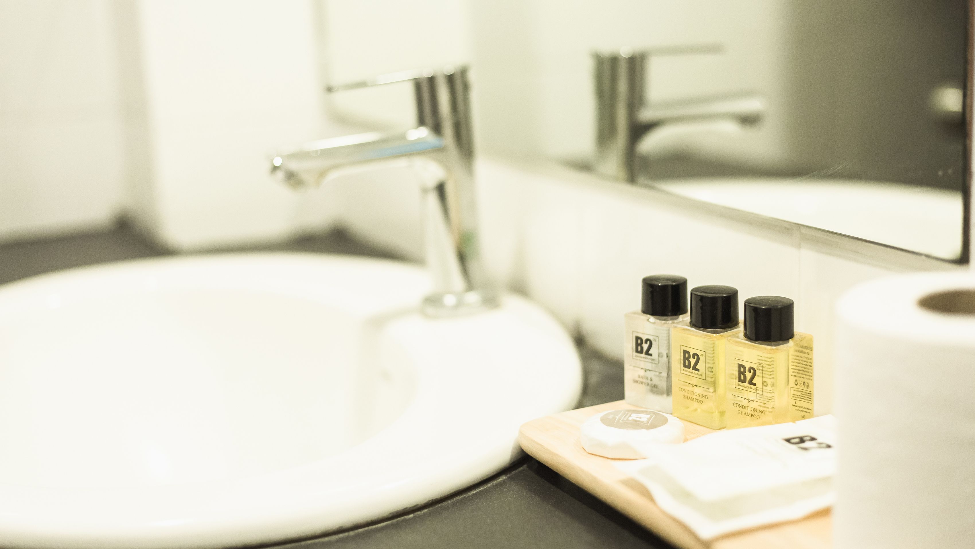 Un lavabo dans une salle de bain, avec un miroir au-dessus, un flacon de shampoing et un rouleau de papier toilette.