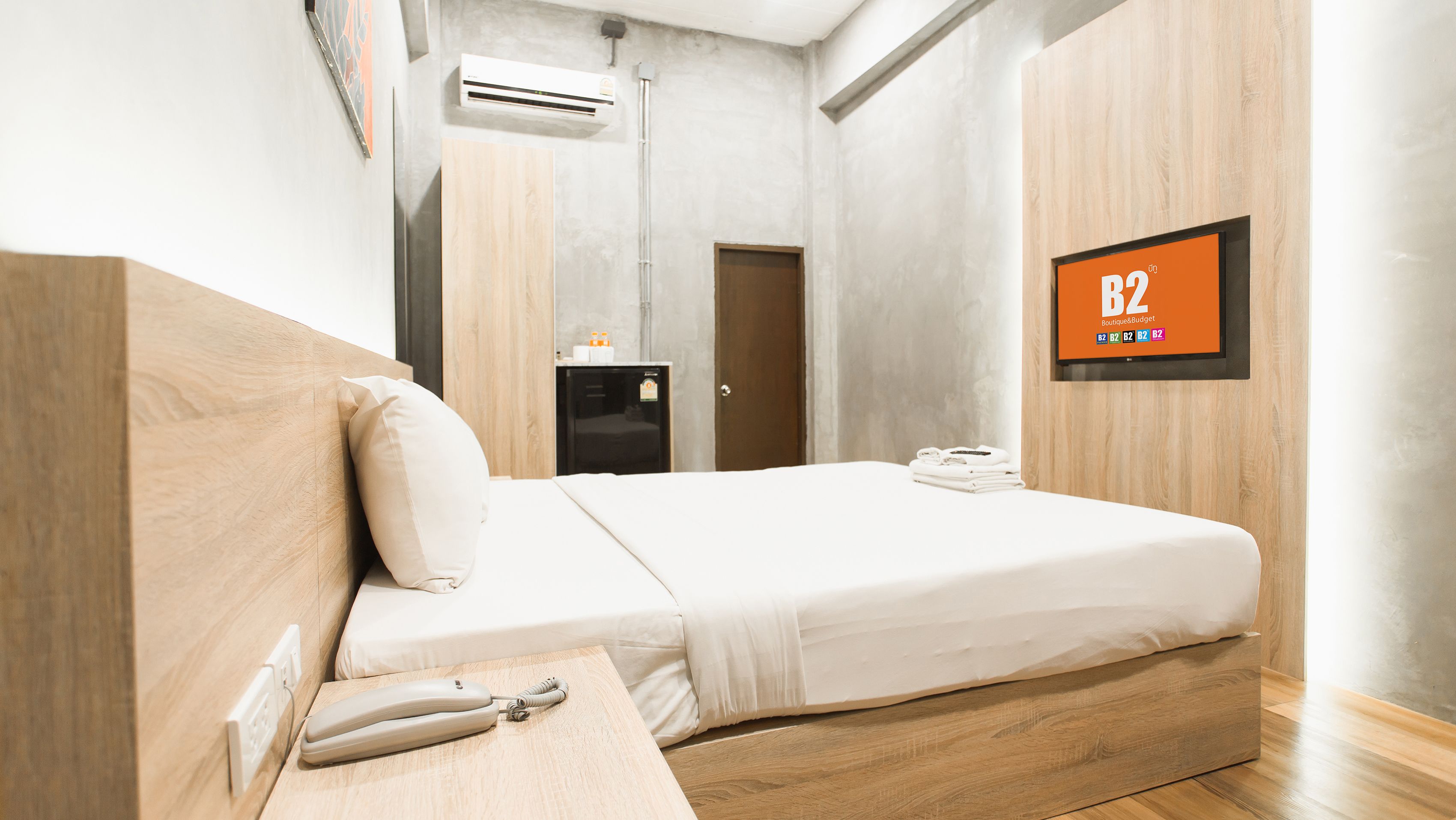 Quarto de hotel moderno com cabeceira de madeira, cama com lençóis brancos, mesa de cabeceira com telefone, TV na parede e unidade de ar condicionado no teto.