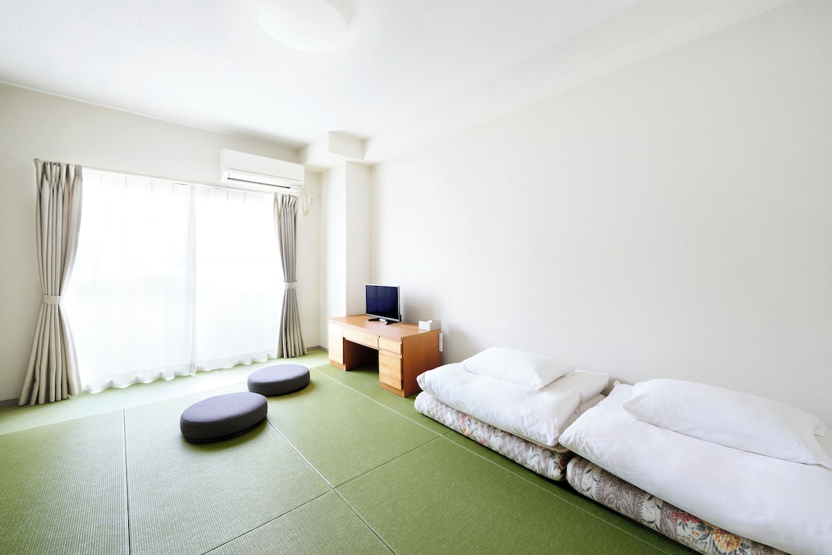 A minimalist hotel room with a green tatami mat floor, a bed with white bedding, a small wooden desk with a television, and a window with curtains.